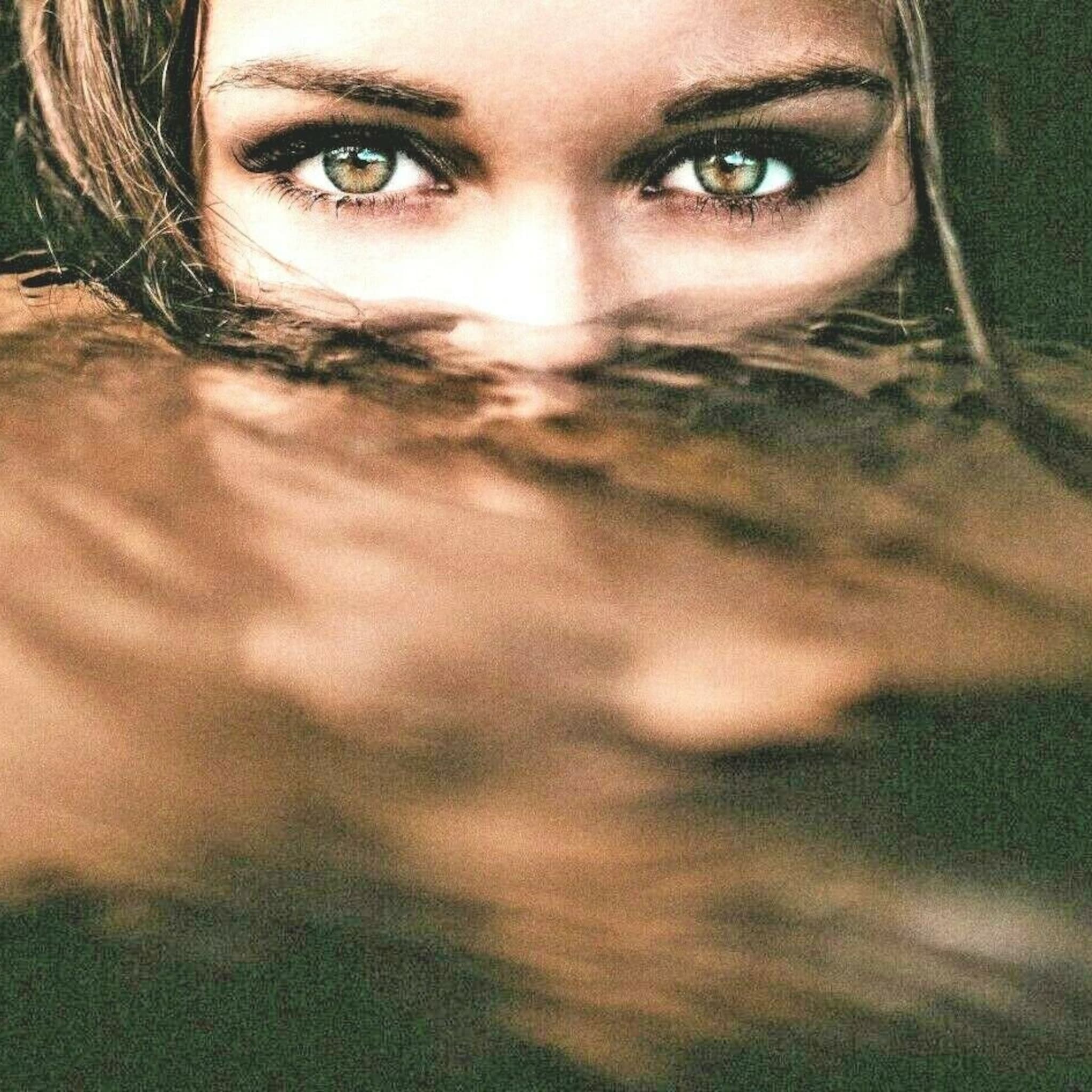 Eyes with green irises, dark eyeliner, and brown hair partially submerged in water