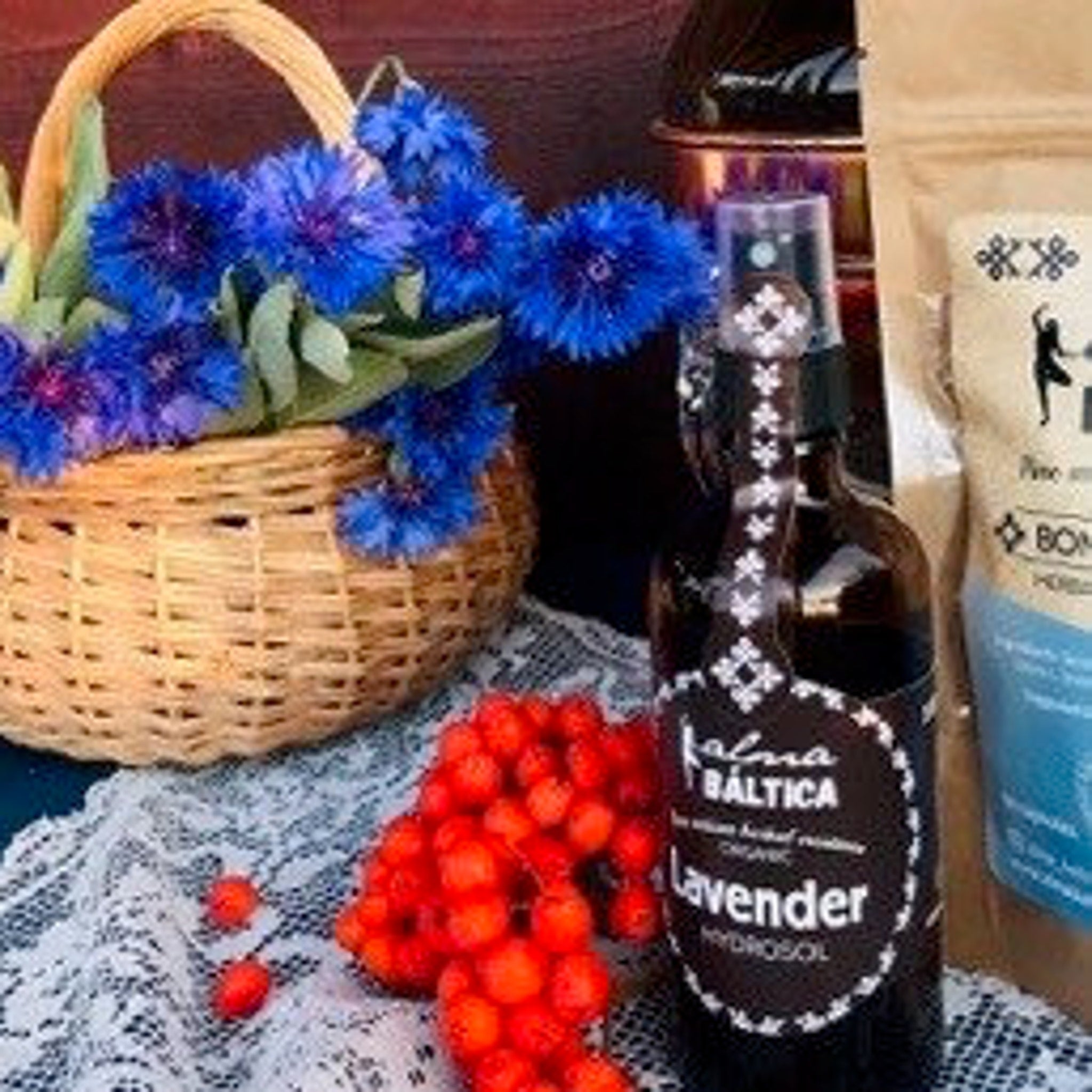 Blue flowers in wicker basket with red berries on white lace, Kalma Baltica lavender hydrosol bottle