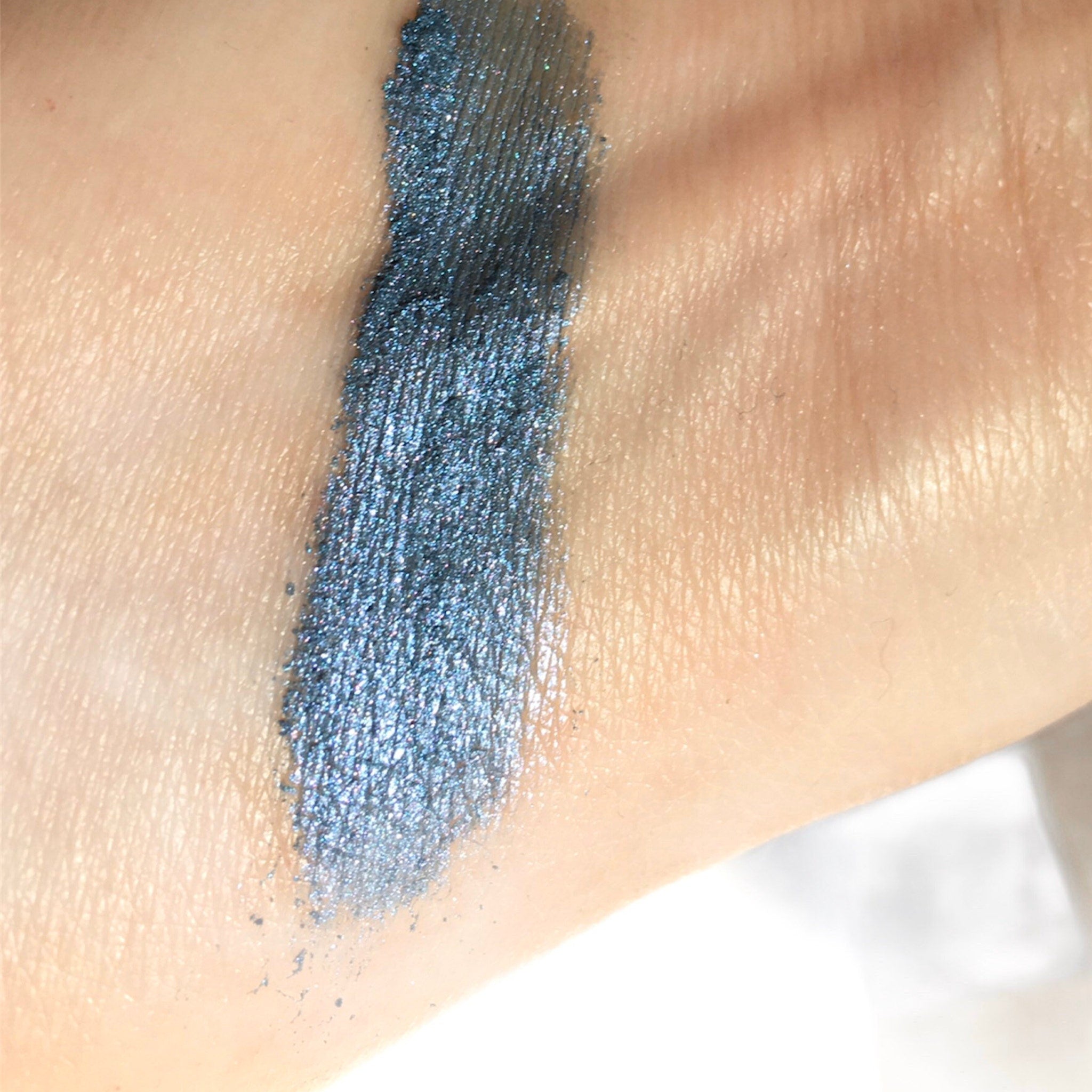Eyeshadow swatch of blue-green metallic pigment on skin, showing texture and color
