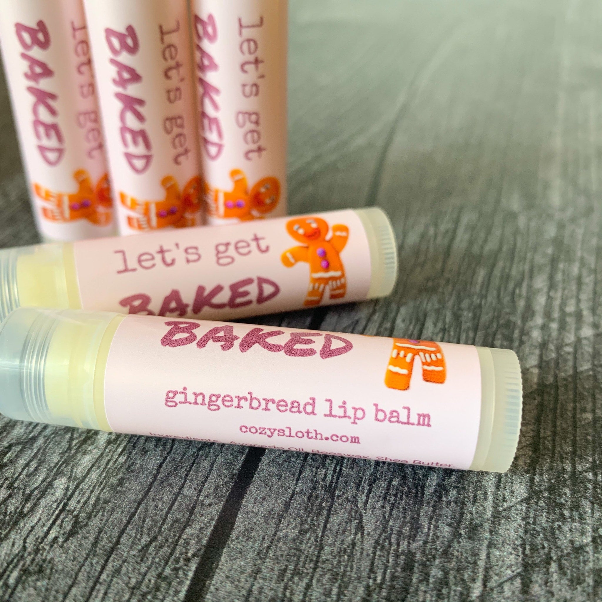 Lip balm tubes with pink labels, orange gingerbread man, and "BAKED" text, on a wooden surface