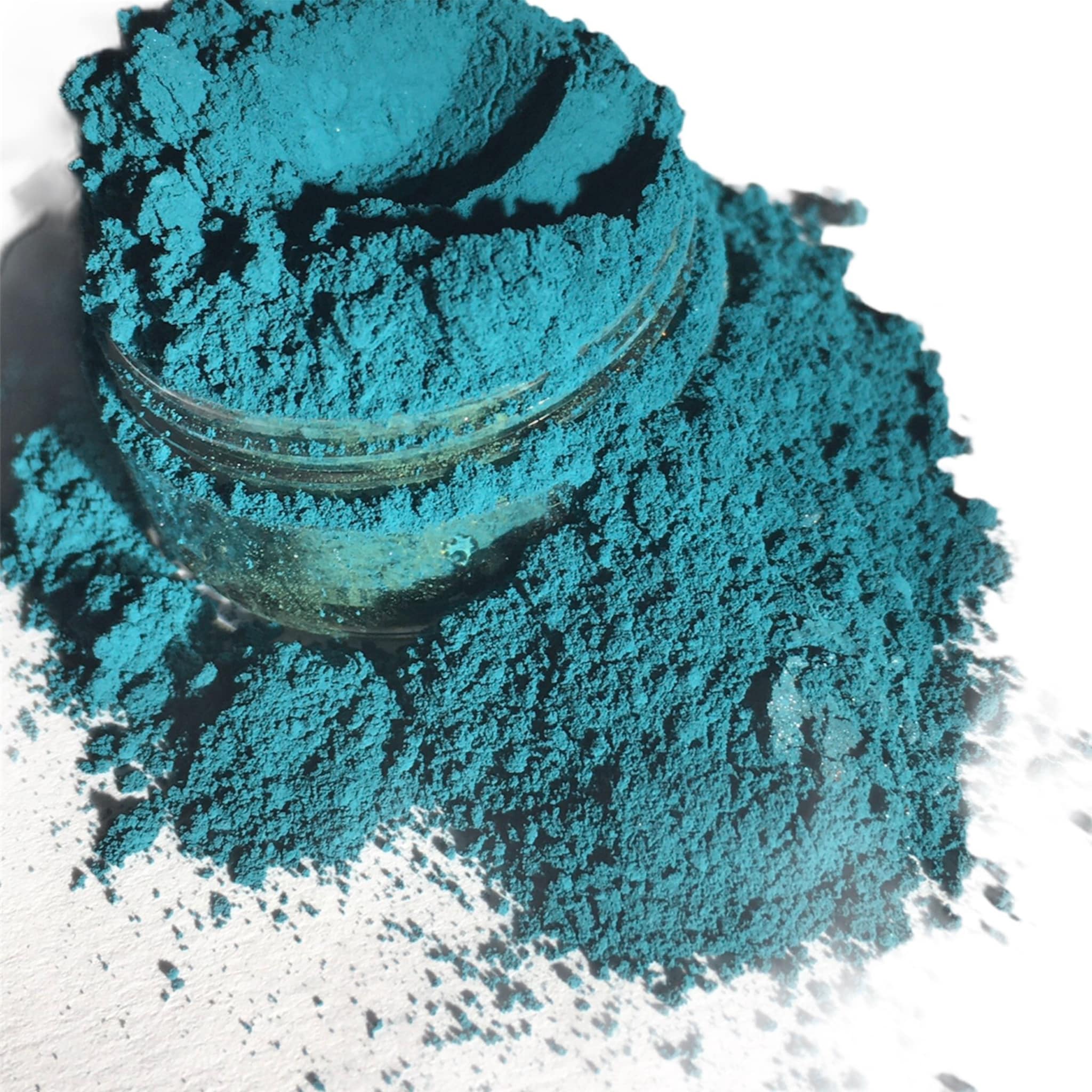 Teal powder pigment with a textured appearance in a glass jar and scattered on white surface