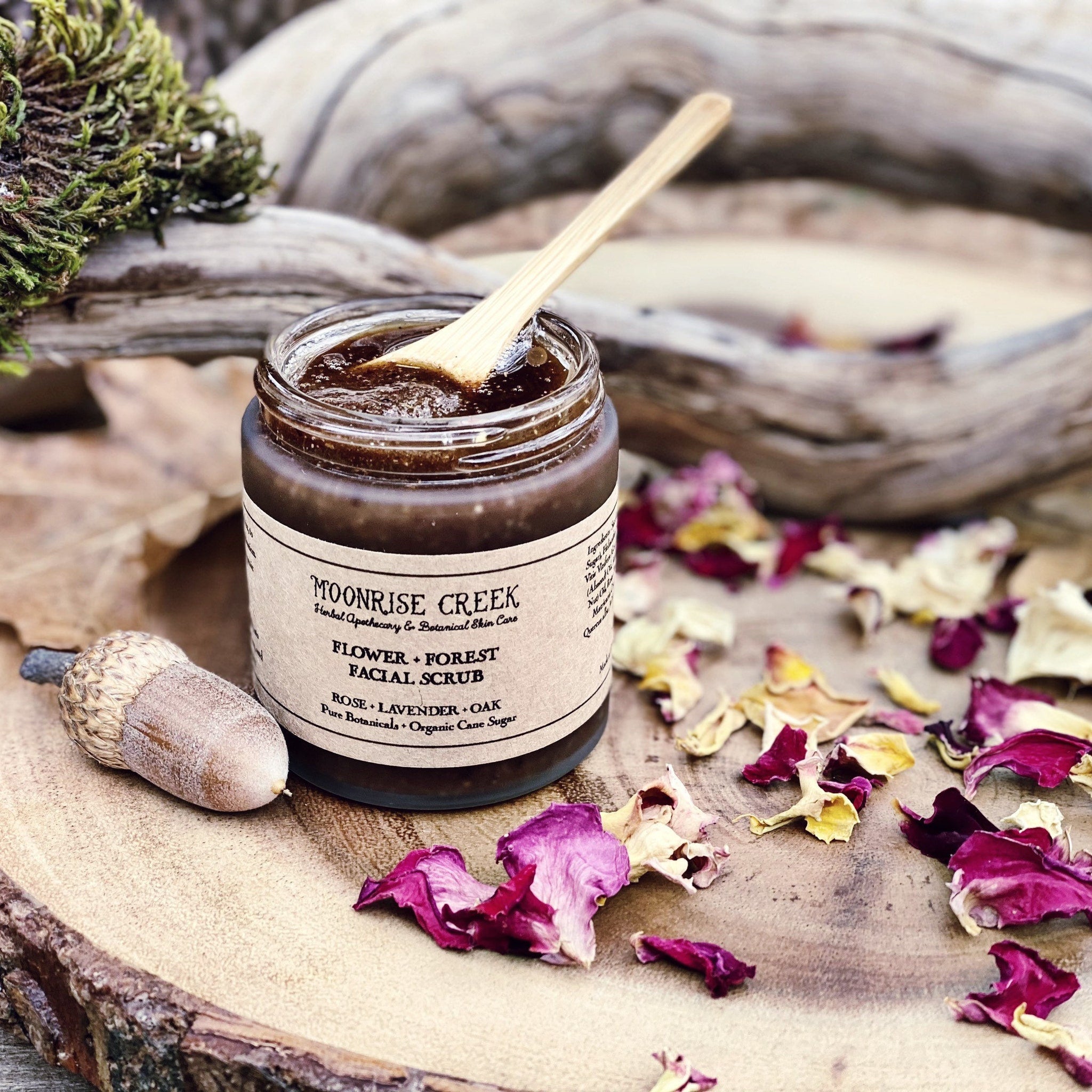 Facial scrub in glass jar with wooden spoon, surrounded by flower petals and acorn on wood surface