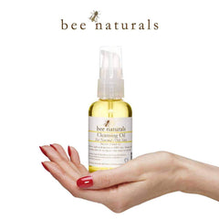 Cleansing oil bottle with yellow liquid, clear pump, and white label held in hand with red nails, Bee Naturals brand visible