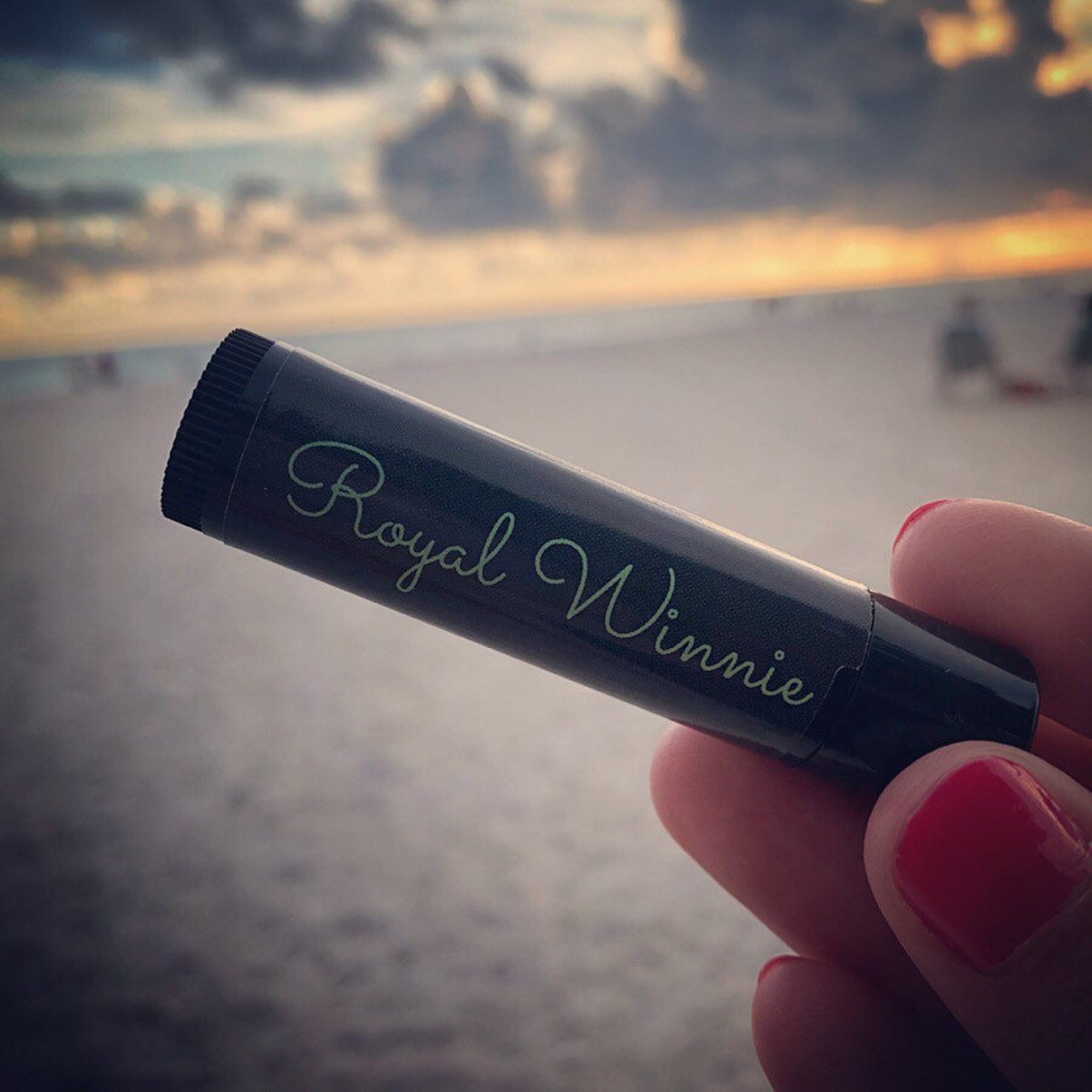 Lip balm, black tube with green Royal Winnie text, held in hand with red nails