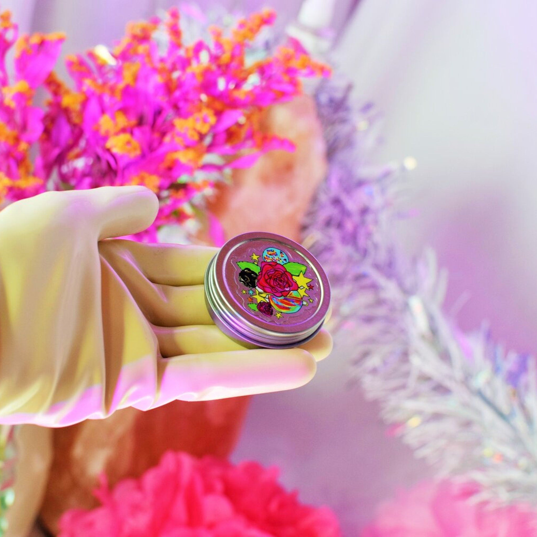 Round metal container with floral illustration, held in a yellow gloved hand with pink and purple background