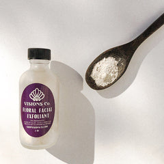 Facial exfoliant bottle with purple label, white powder, and wooden spoon on white surface