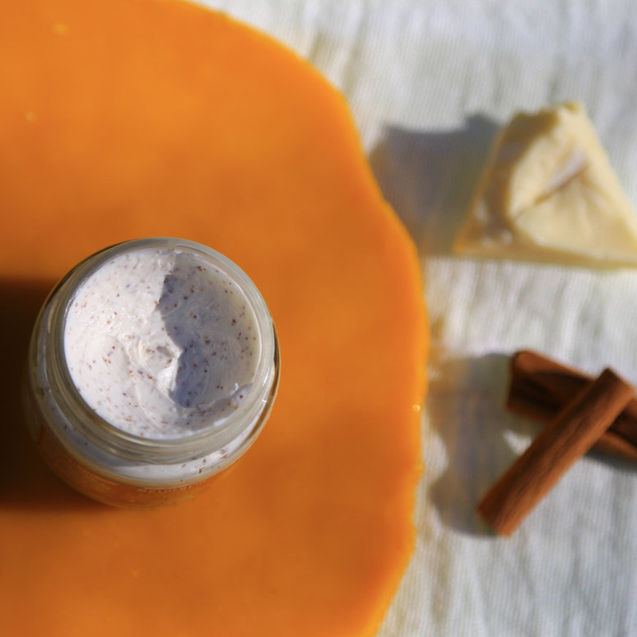 Body scrub in glass jar with white and brown speckled cream on orange surface with cinnamon sticks