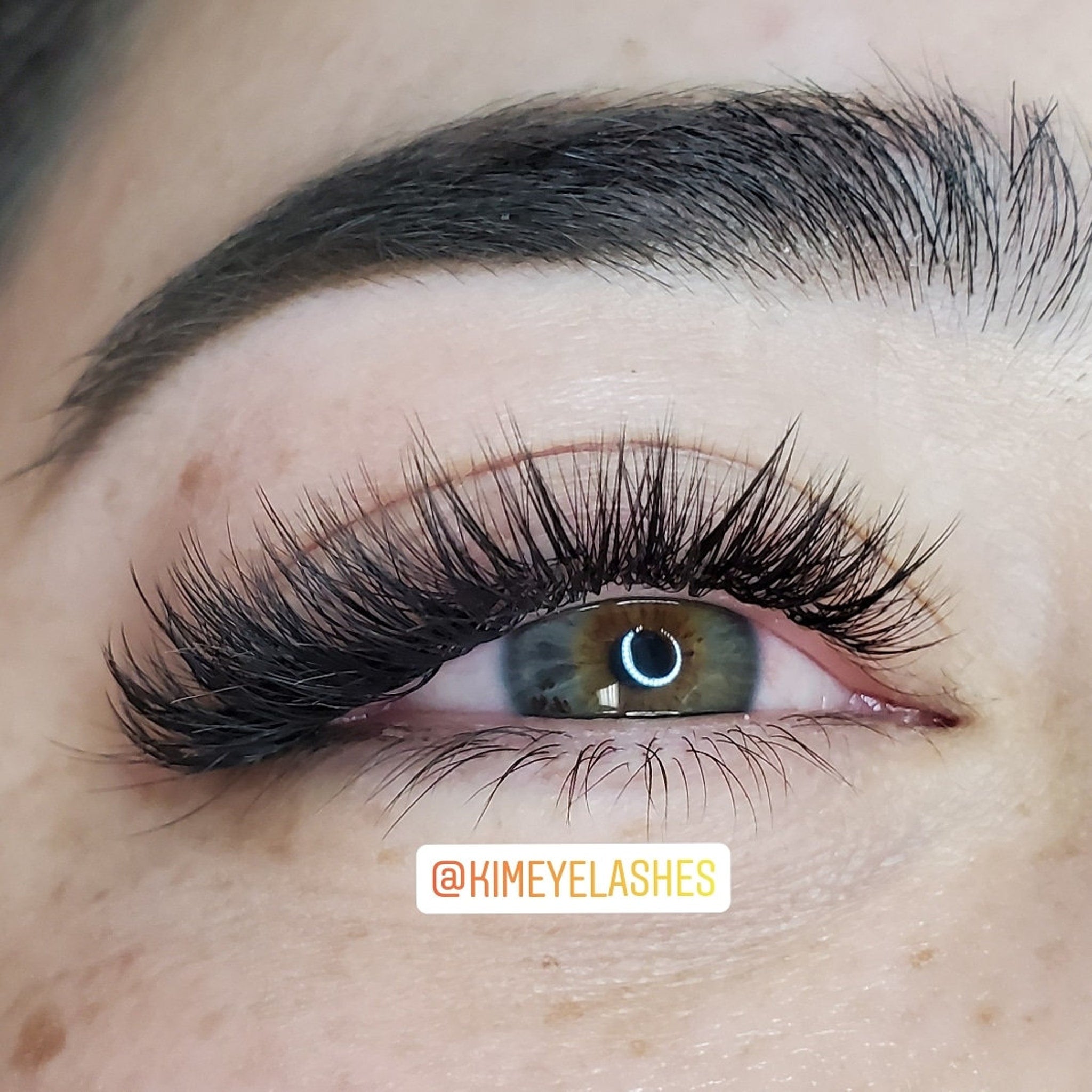 Eyelash extensions with black lashes, brown and green eye, and dark eyebrow, @KIMEYELASHES logo visible