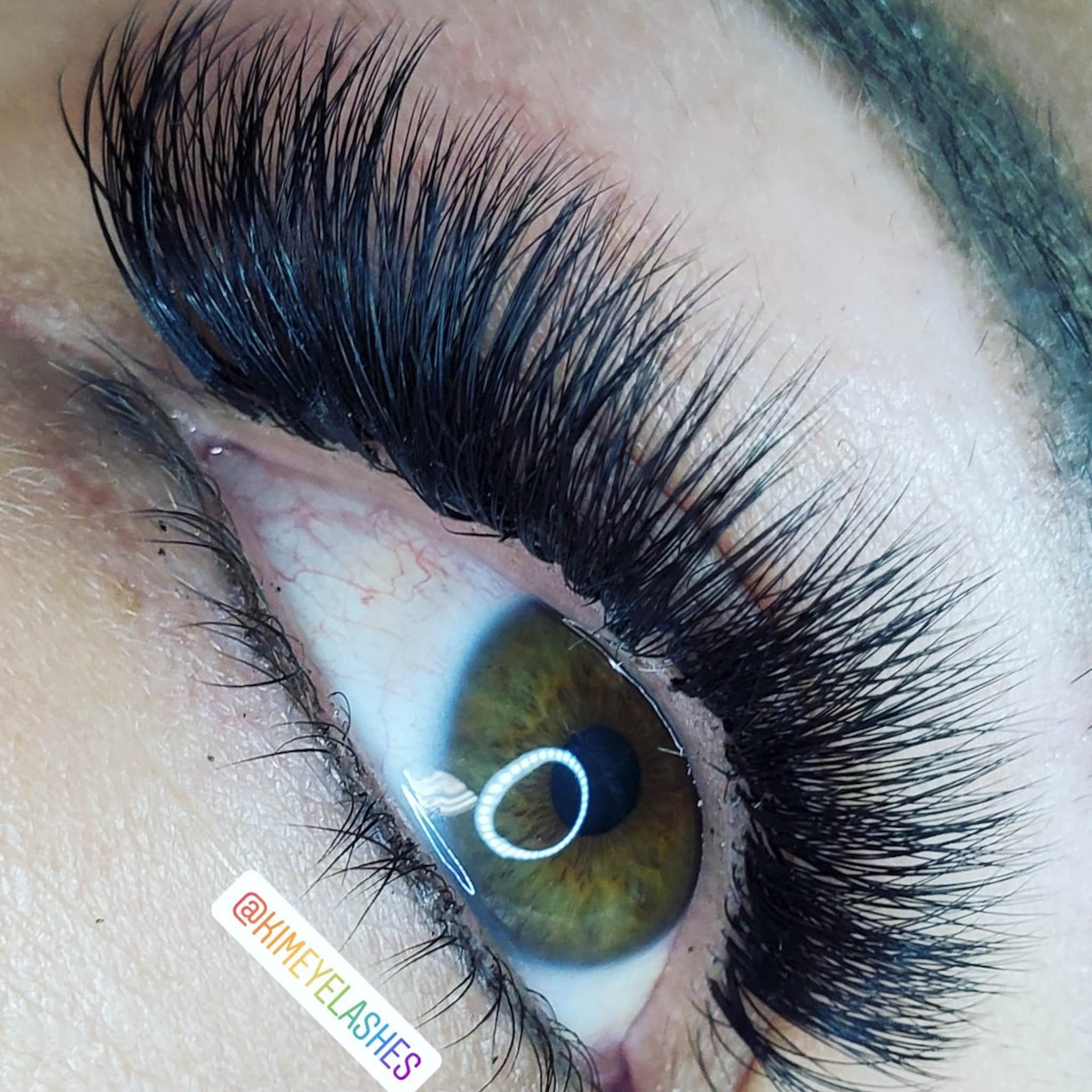 Eyelash extensions with black, long, and curved synthetic fibers, visible around an eye with brand name @KIMEYELASHES