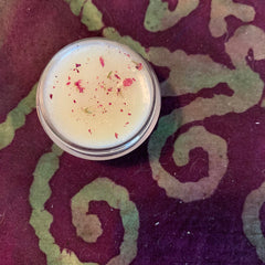 Lip balm in clear jar with cream color and red flower petals, on patterned purple fabric
