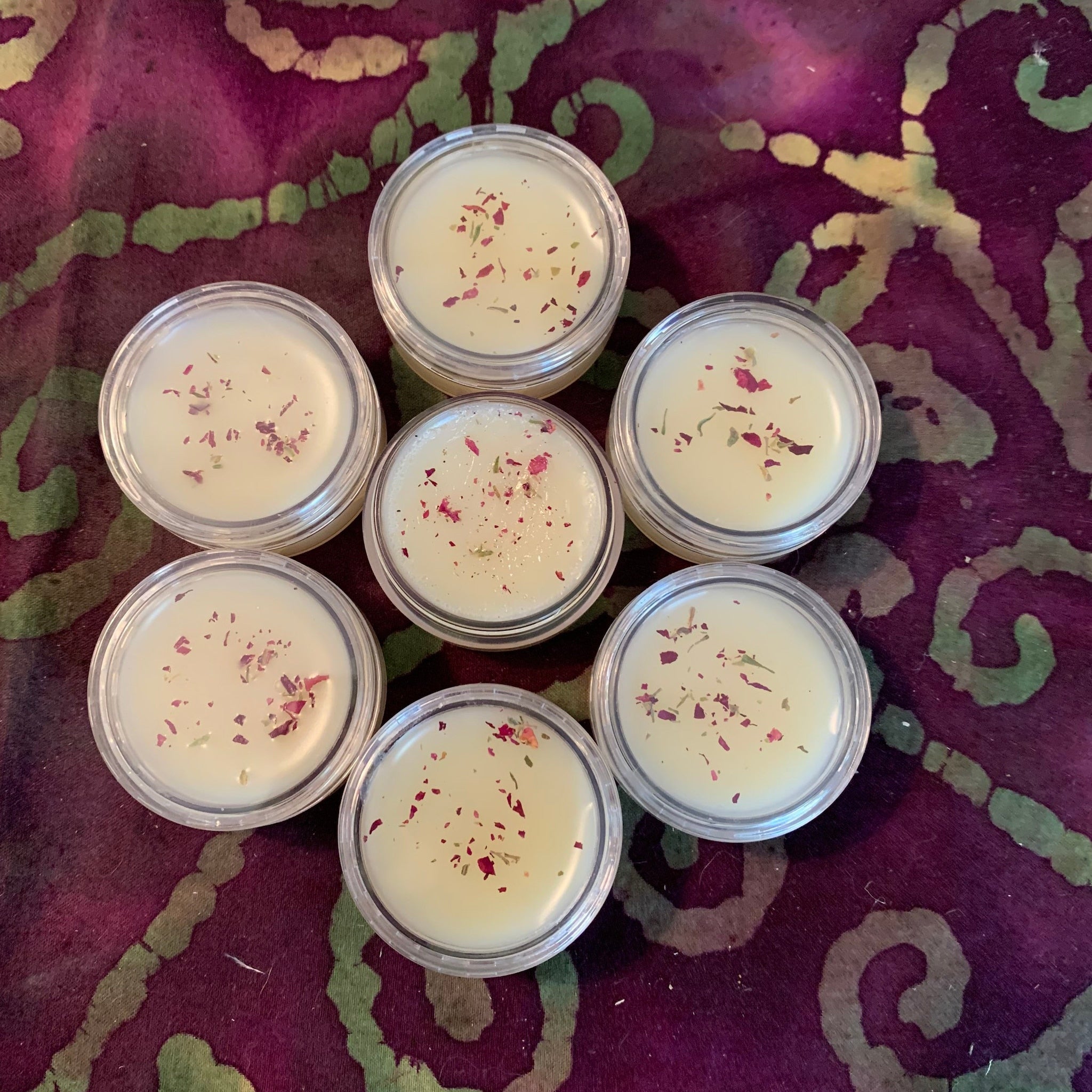 Lip balm in clear plastic containers with light yellow balm and dried flower petals on purple cloth