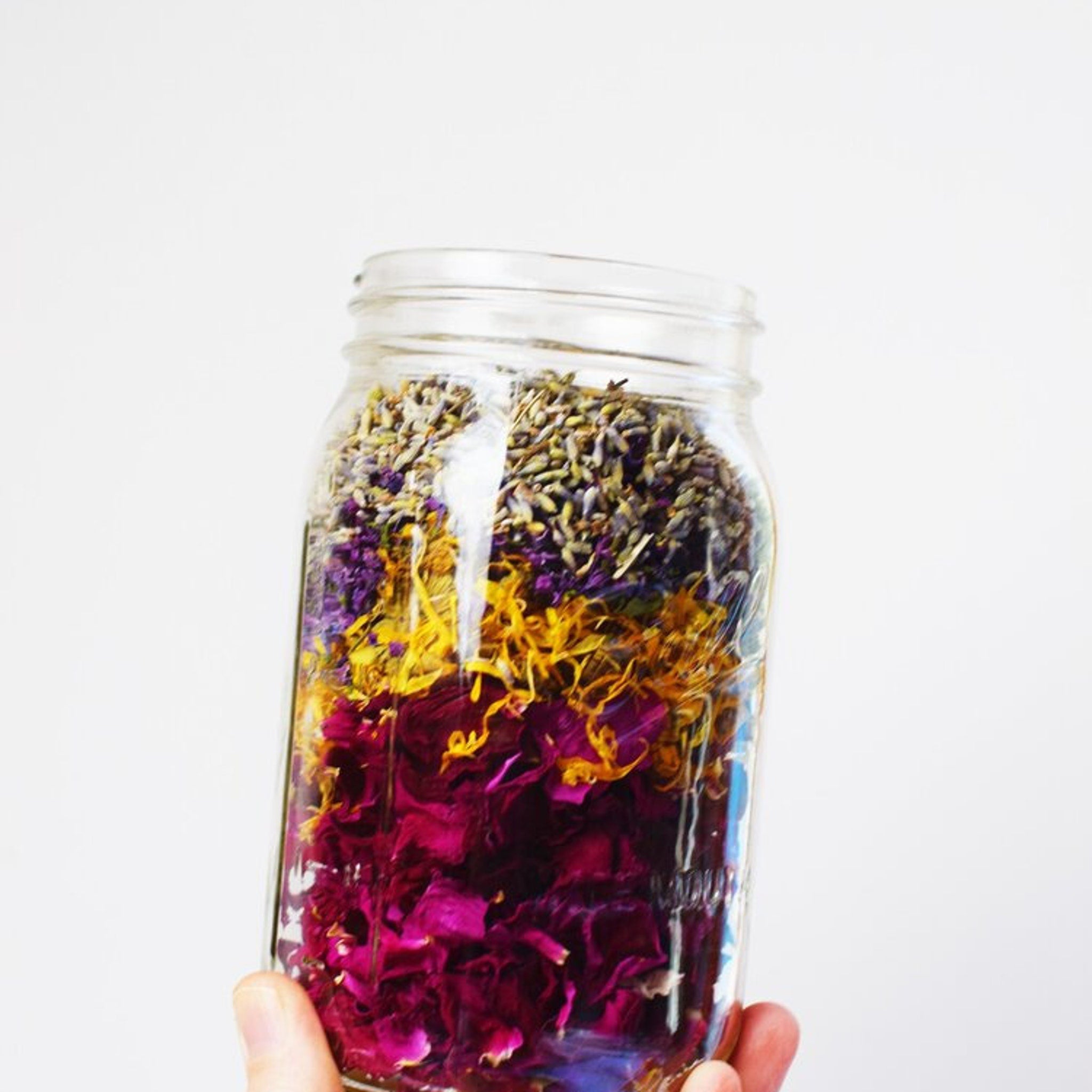 Dried flower arrangement in a clear glass jar with layers of purple, yellow, and red petals, held by a hand