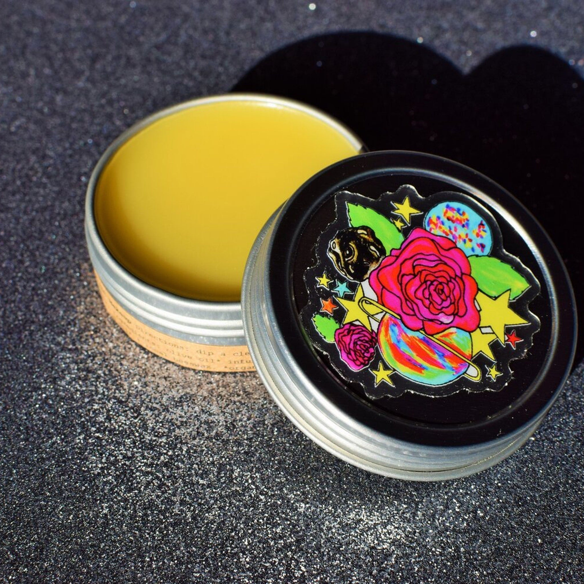 Lip balm in silver tin with yellow balm, black lid with colorful floral design, open