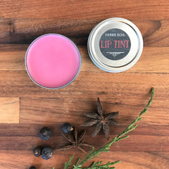 Lip tint in pink color inside a metal tin, Herbs Soil branding visible, on a wooden surface