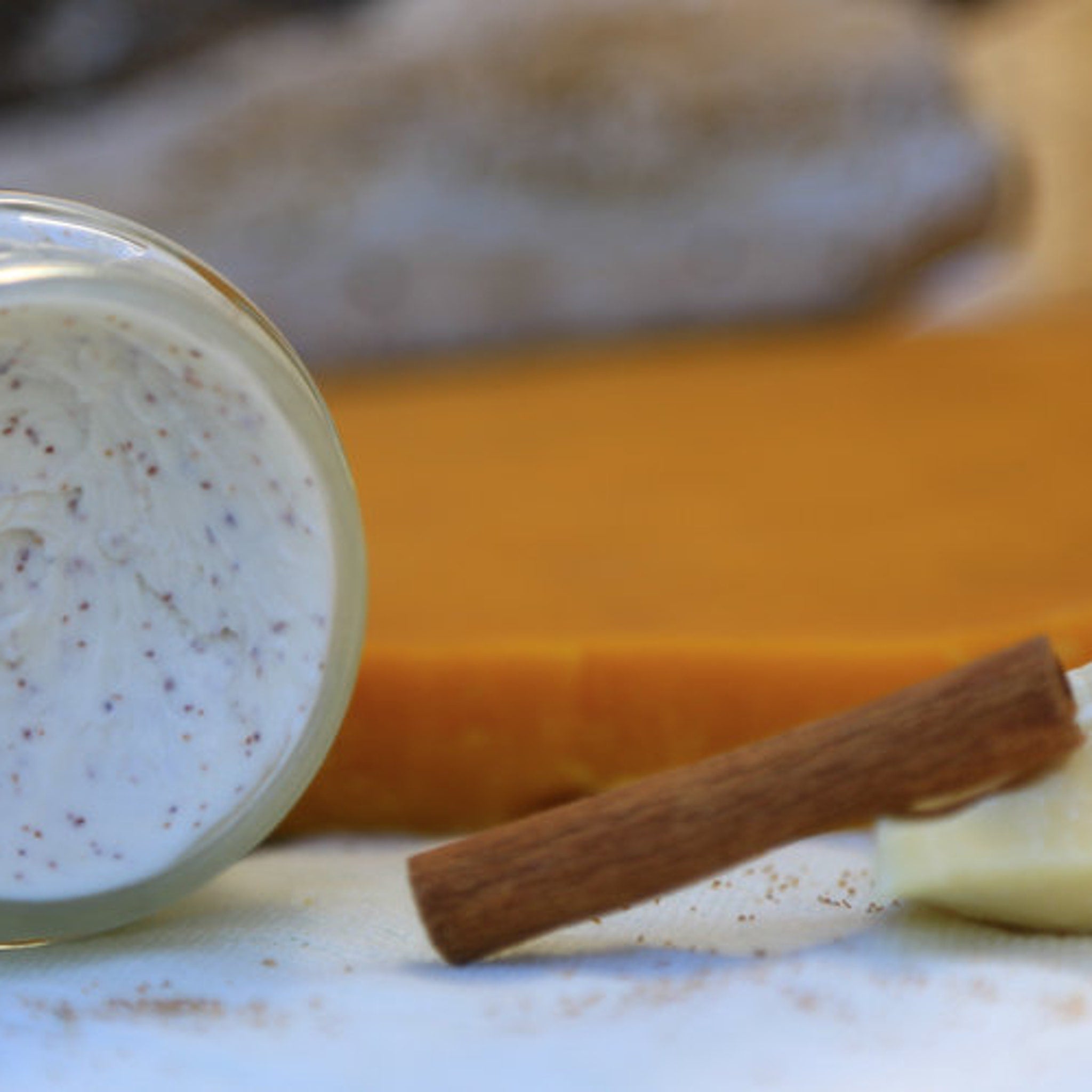 Creamy white lotion with brown specks in a glass jar, with cinnamon stick and orange background.