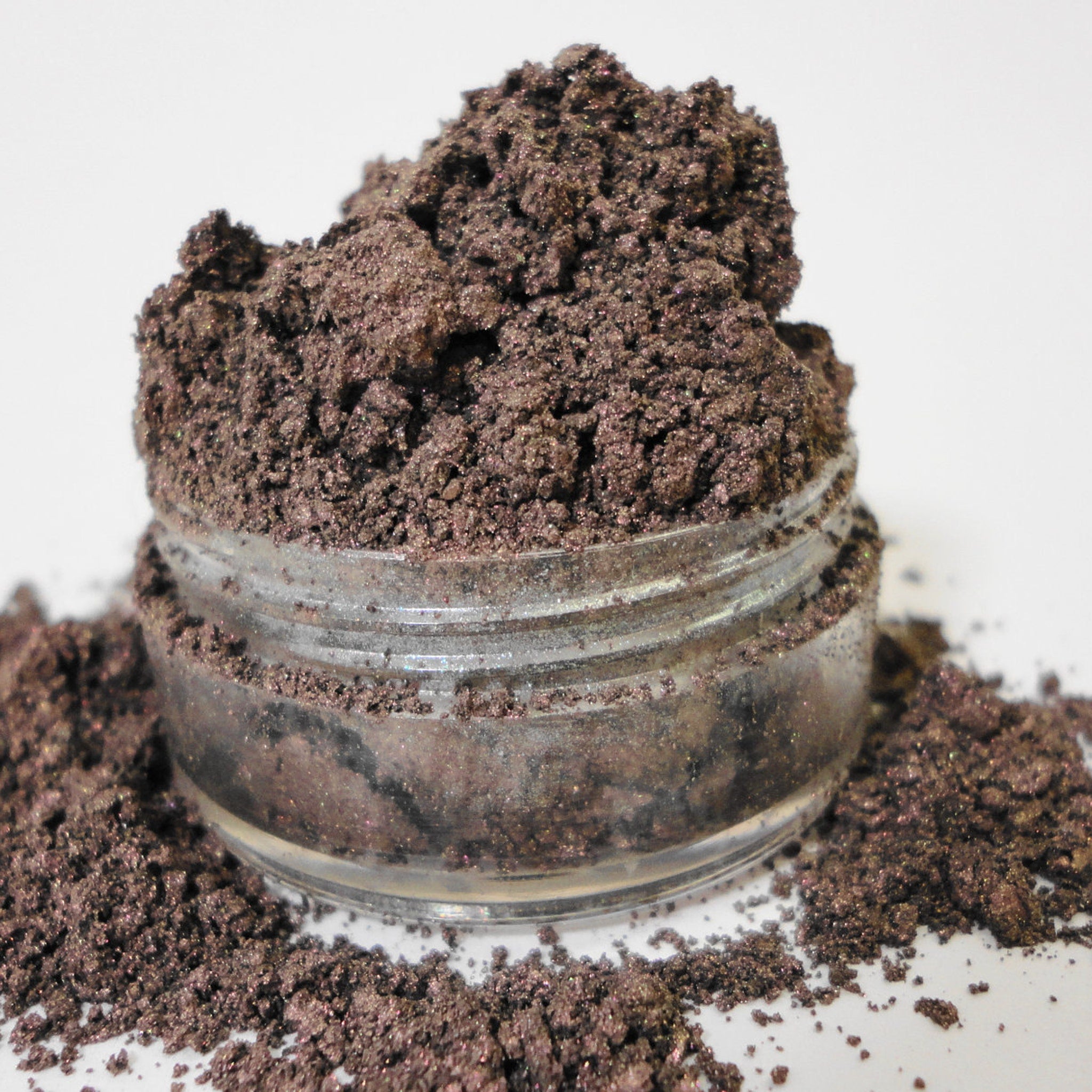 Loose eyeshadow powder, dark brown with shimmer, in a clear glass jar with spilled powder