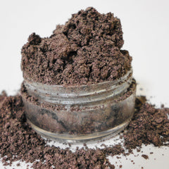 Loose eyeshadow powder, dark brown with shimmer, in a clear glass jar with spilled powder