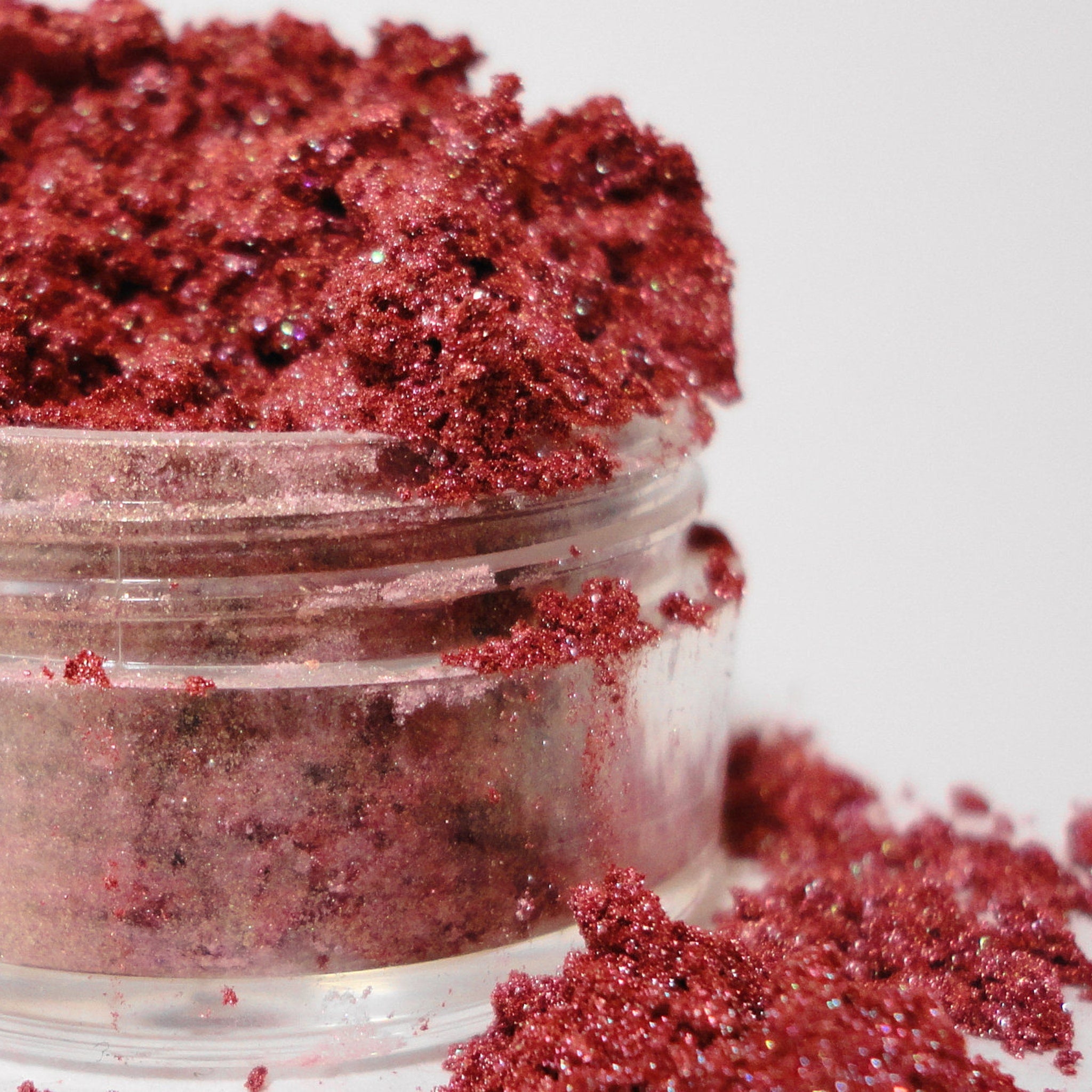 Red glitter eyeshadow in a clear plastic container with scattered particles on white background
