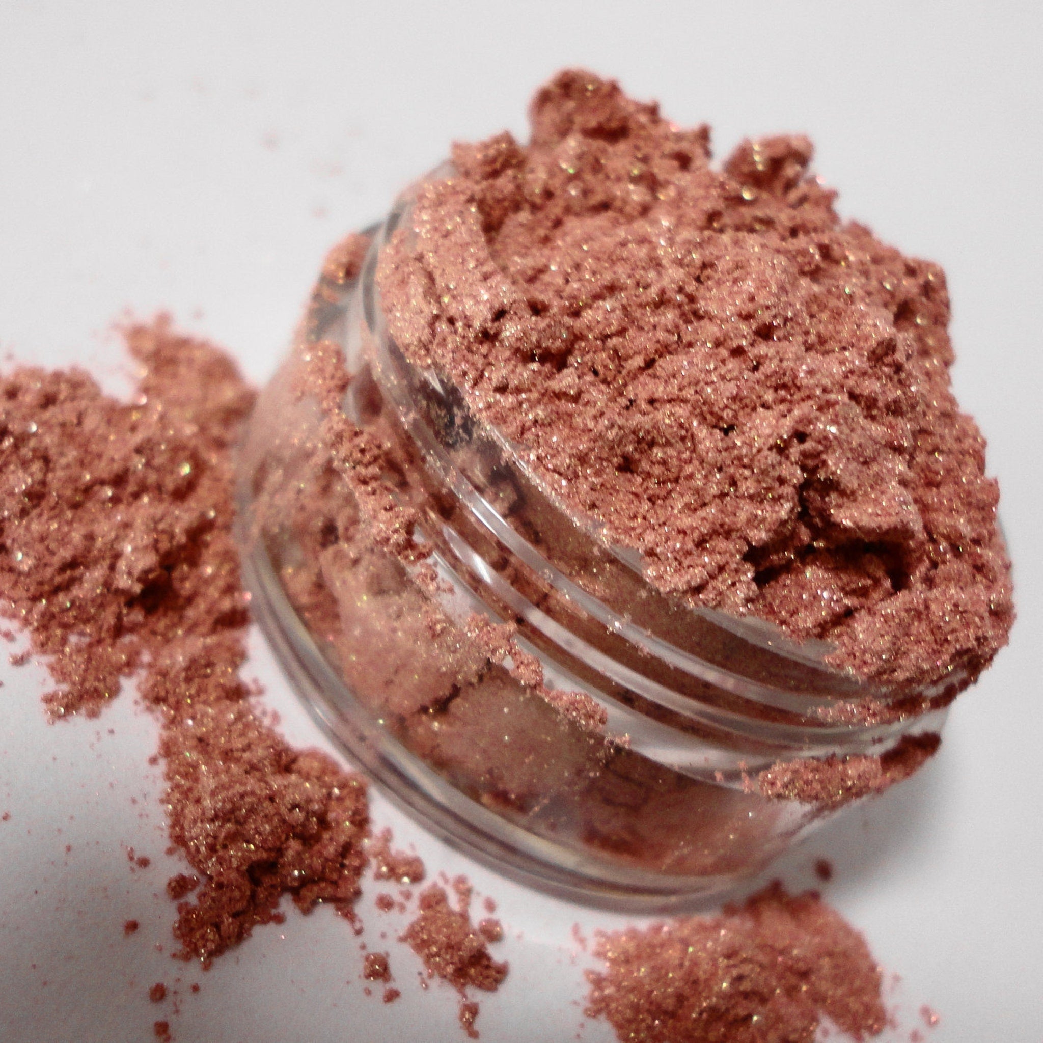Eyeshadow powder in pink with shimmer, in a clear plastic jar, spilled on white surface