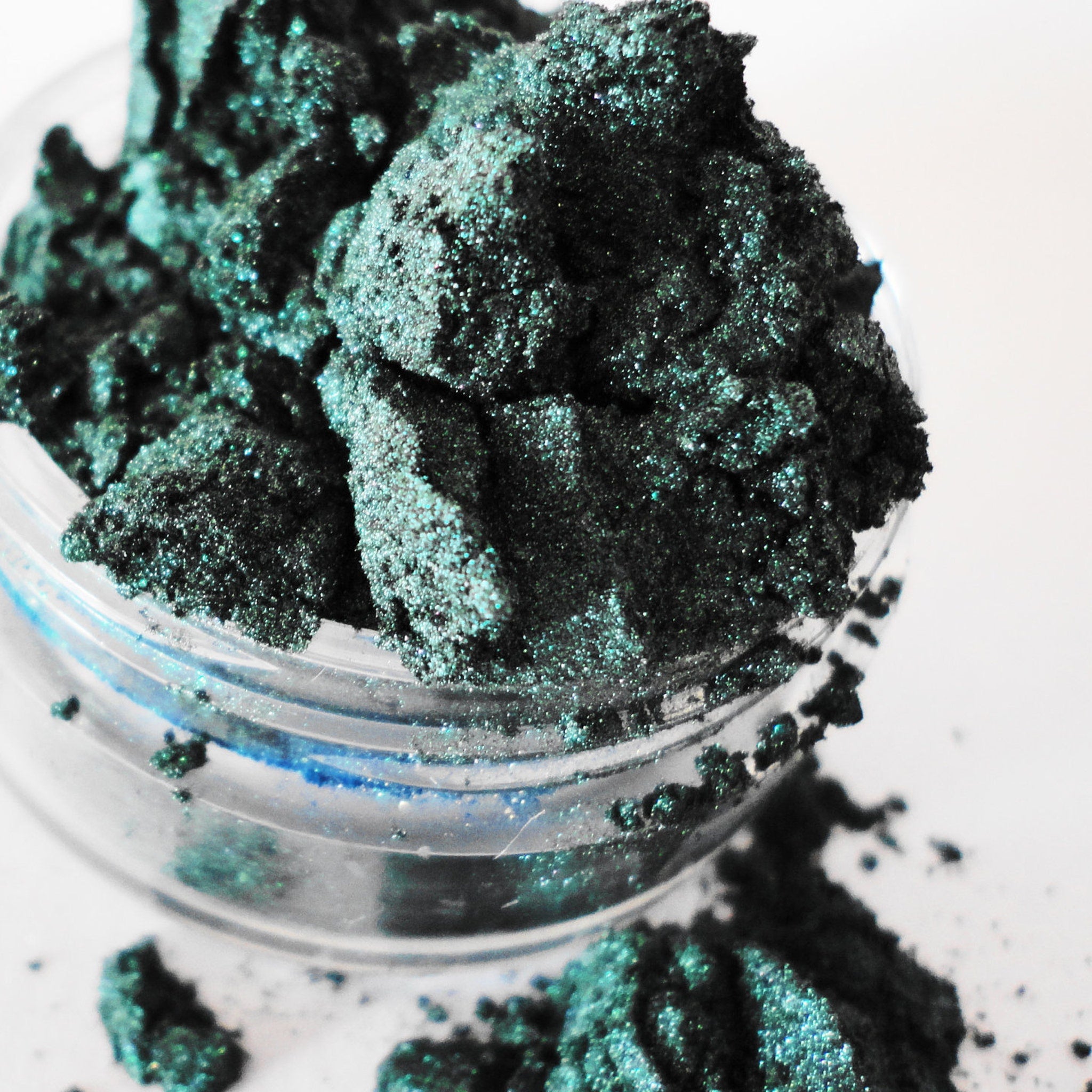 Eyeshadow powder, dark teal green, glittery, in a clear container, on white surface