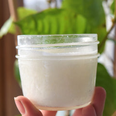 Creamy white jar with clear glass, held by hand, in front of green leaves