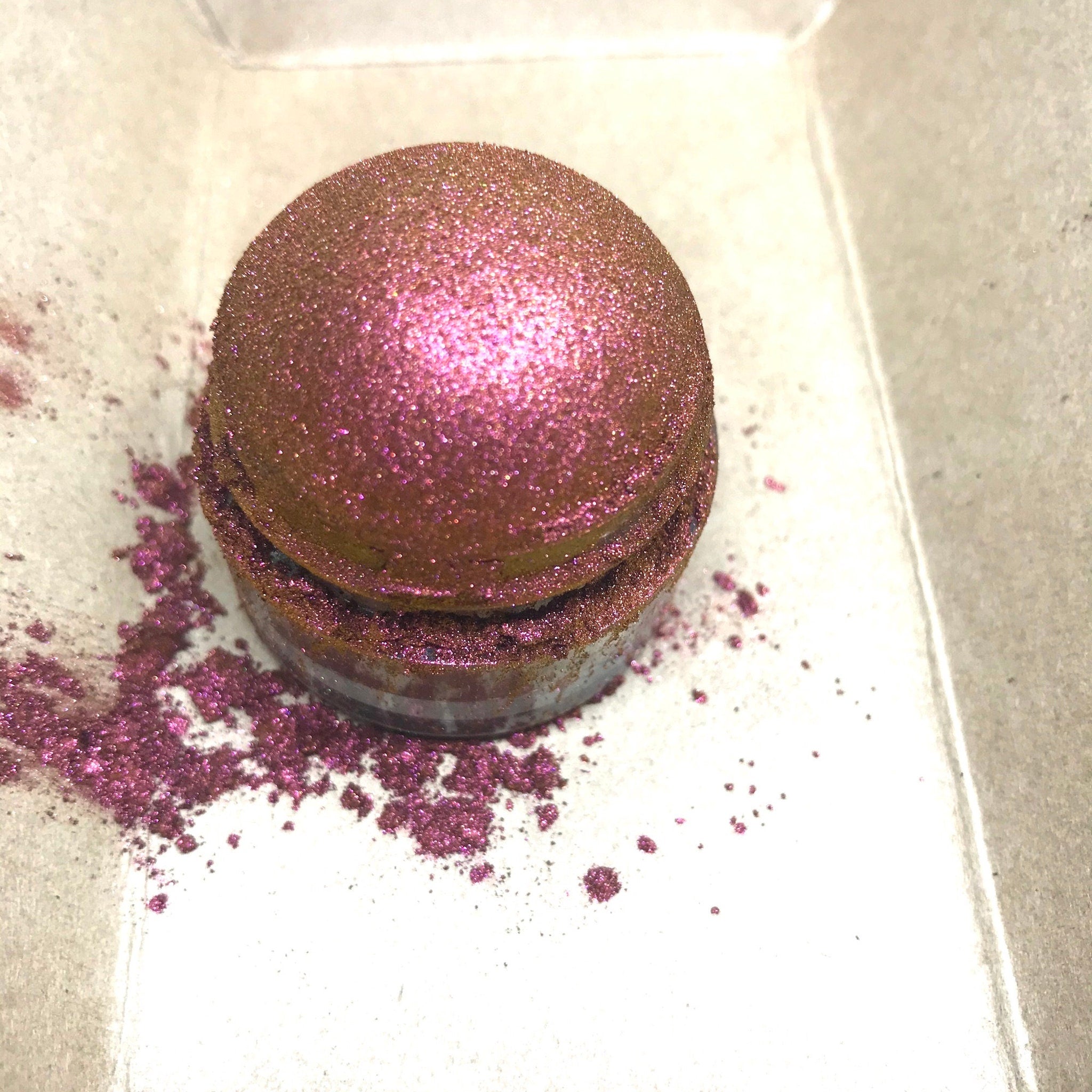 Glitter eyeshadow pot with pink and gold shimmer, spilled powder on white surface