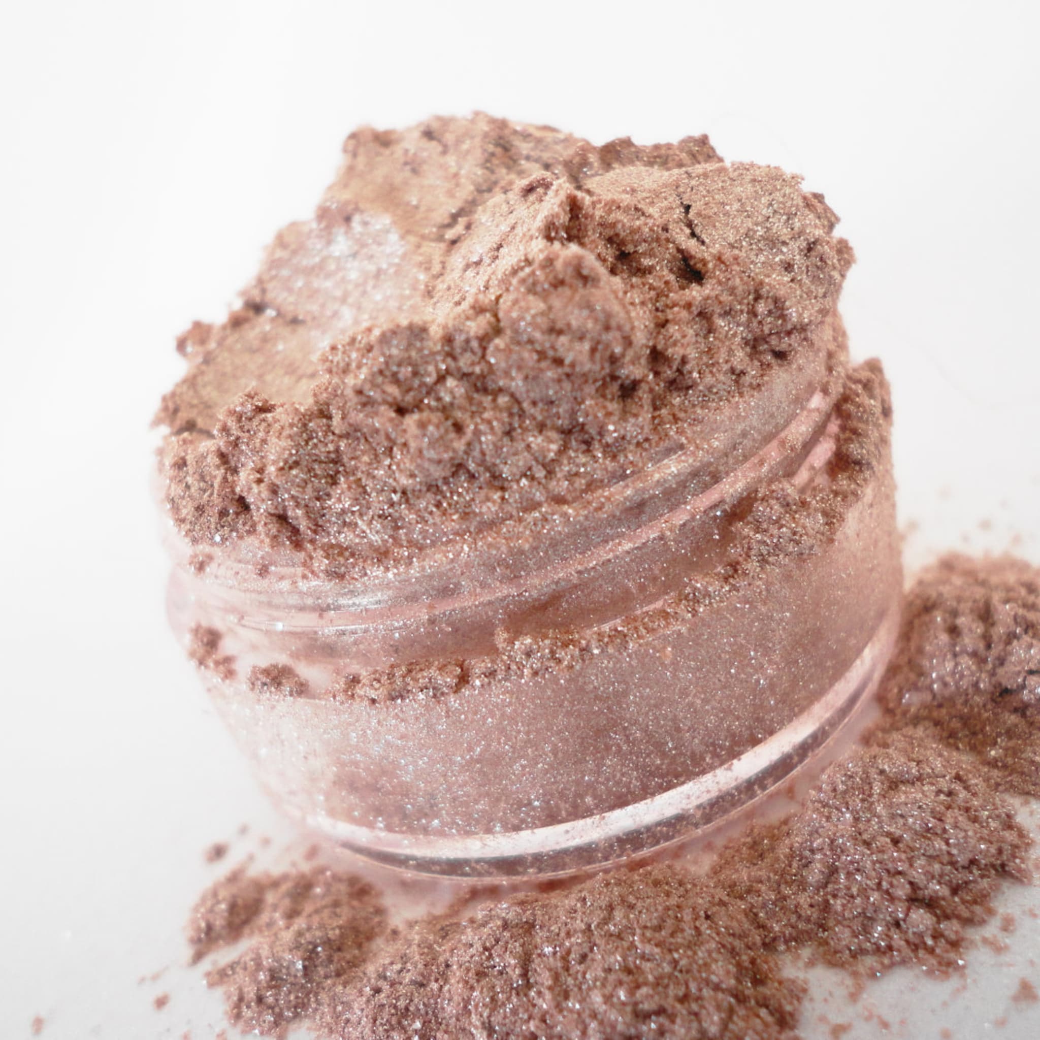 Eyeshadow powder in a shimmering brown color, in a clear jar and spilled on a white surface