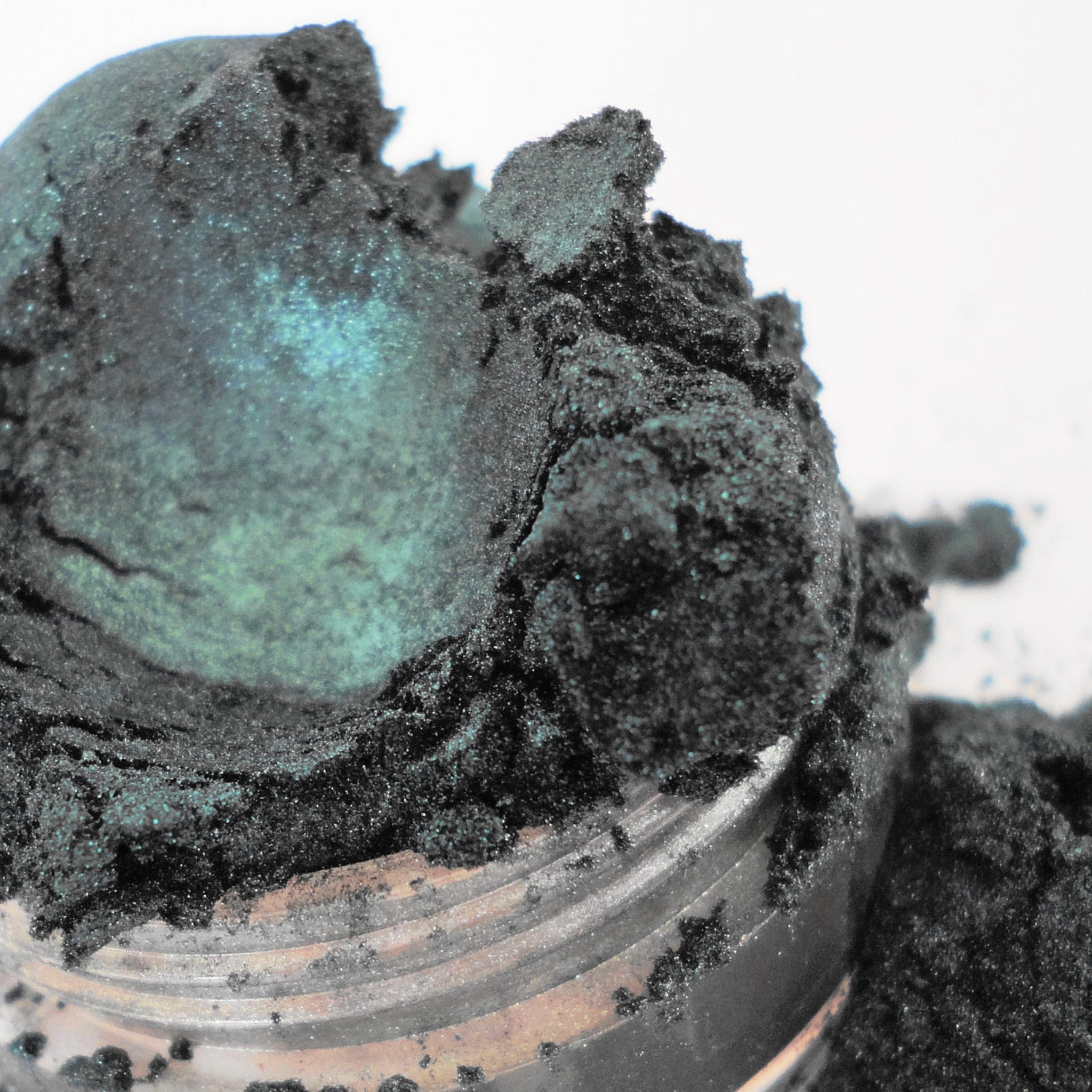Eyeshadow powder in dark teal with glitter and textured appearance, contained in a jar