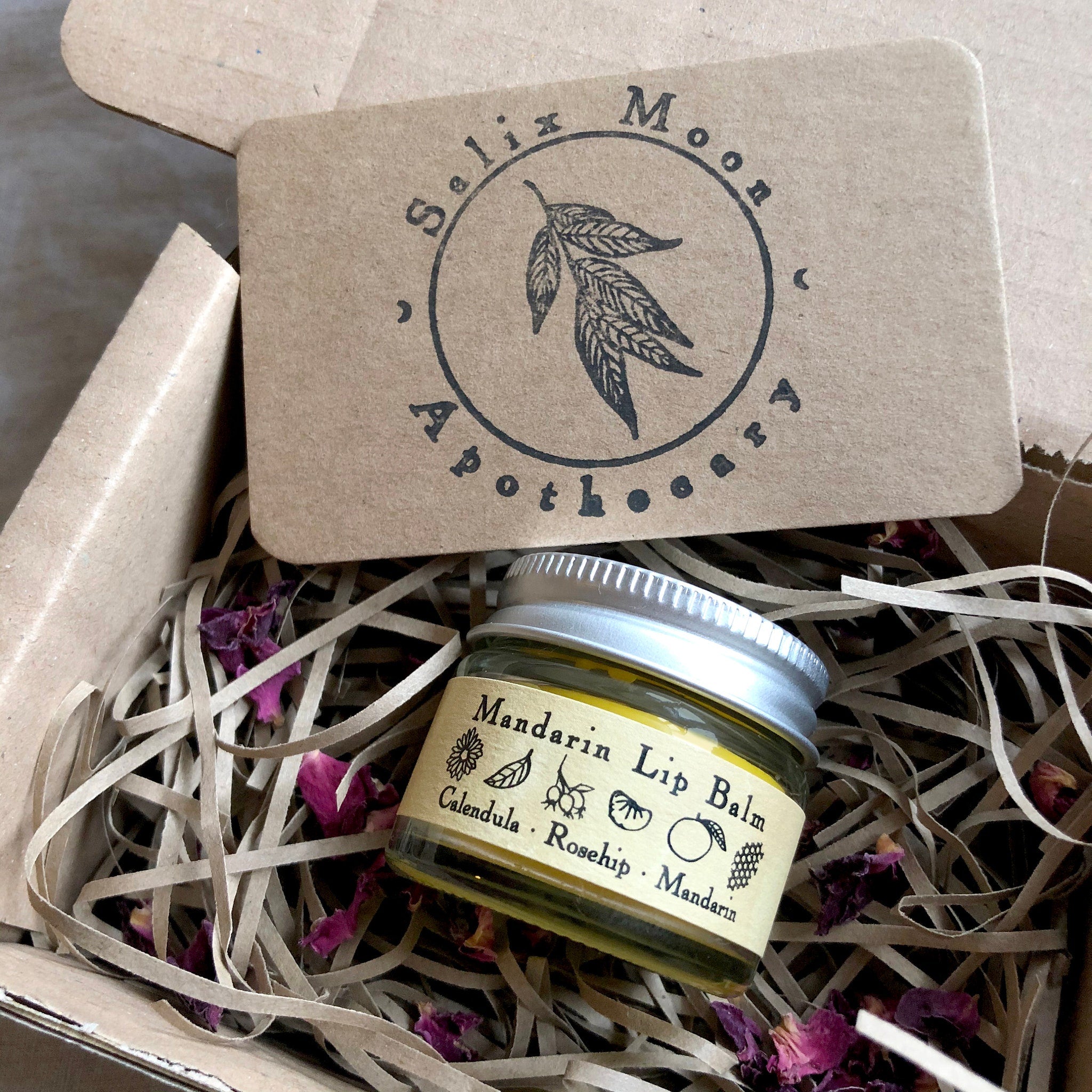 Lip balm jar with yellow liquid, silver lid, and label, in a cardboard box with paper filling and dried flowers