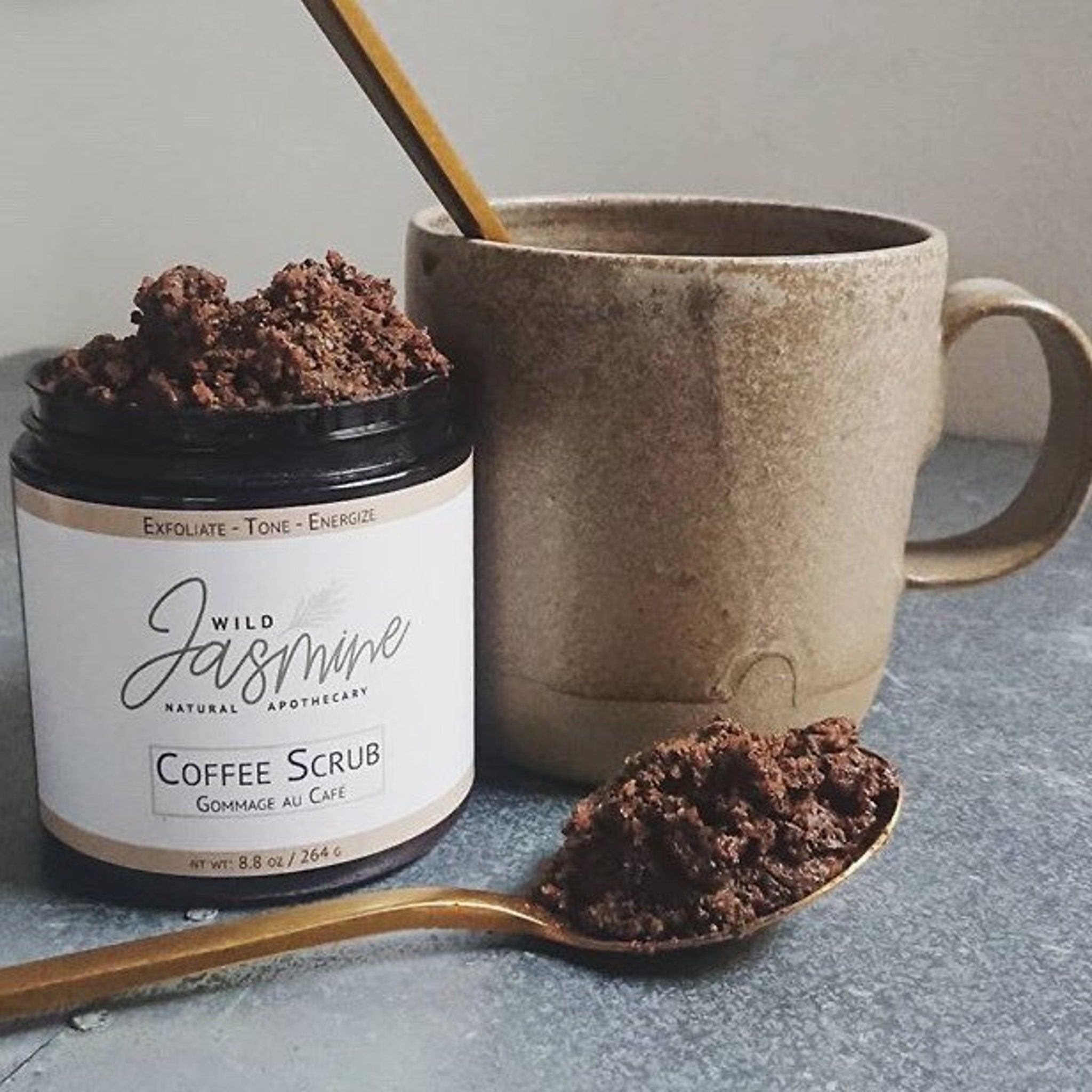 Coffee scrub in a jar with Wild Jasmine label, brown granules, and a ceramic mug with a spoon alongside