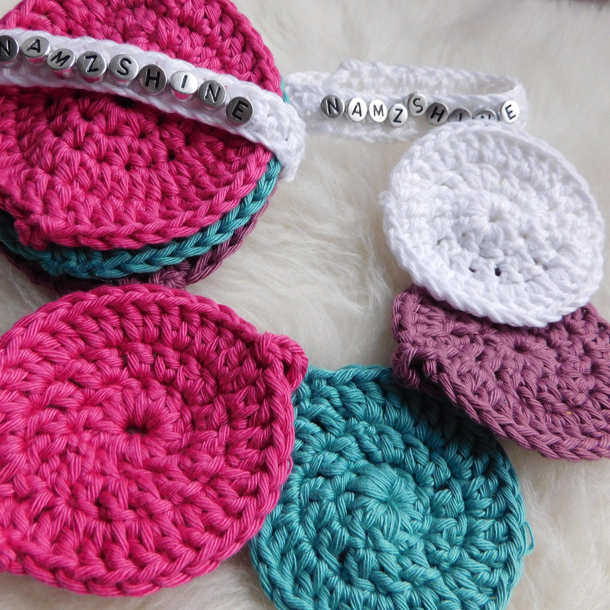 Crochet coasters in pink, white, and teal colors with visible stitching and beaded text details