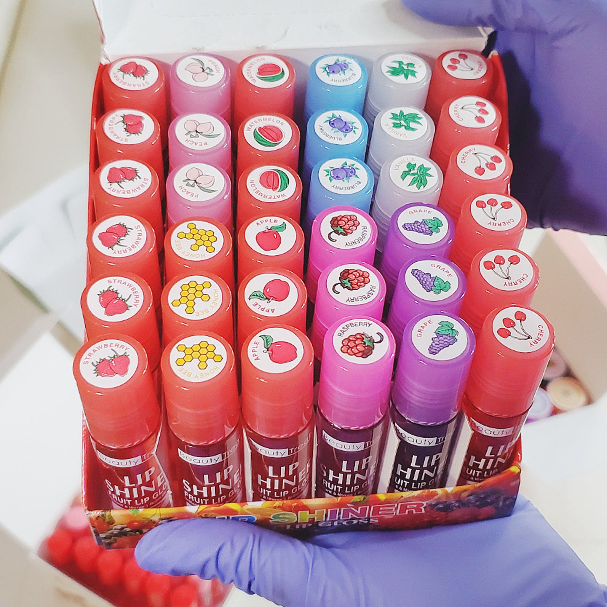 Lip gloss tubes in red, pink, and purple with fruit labels, in a box held by gloved hands