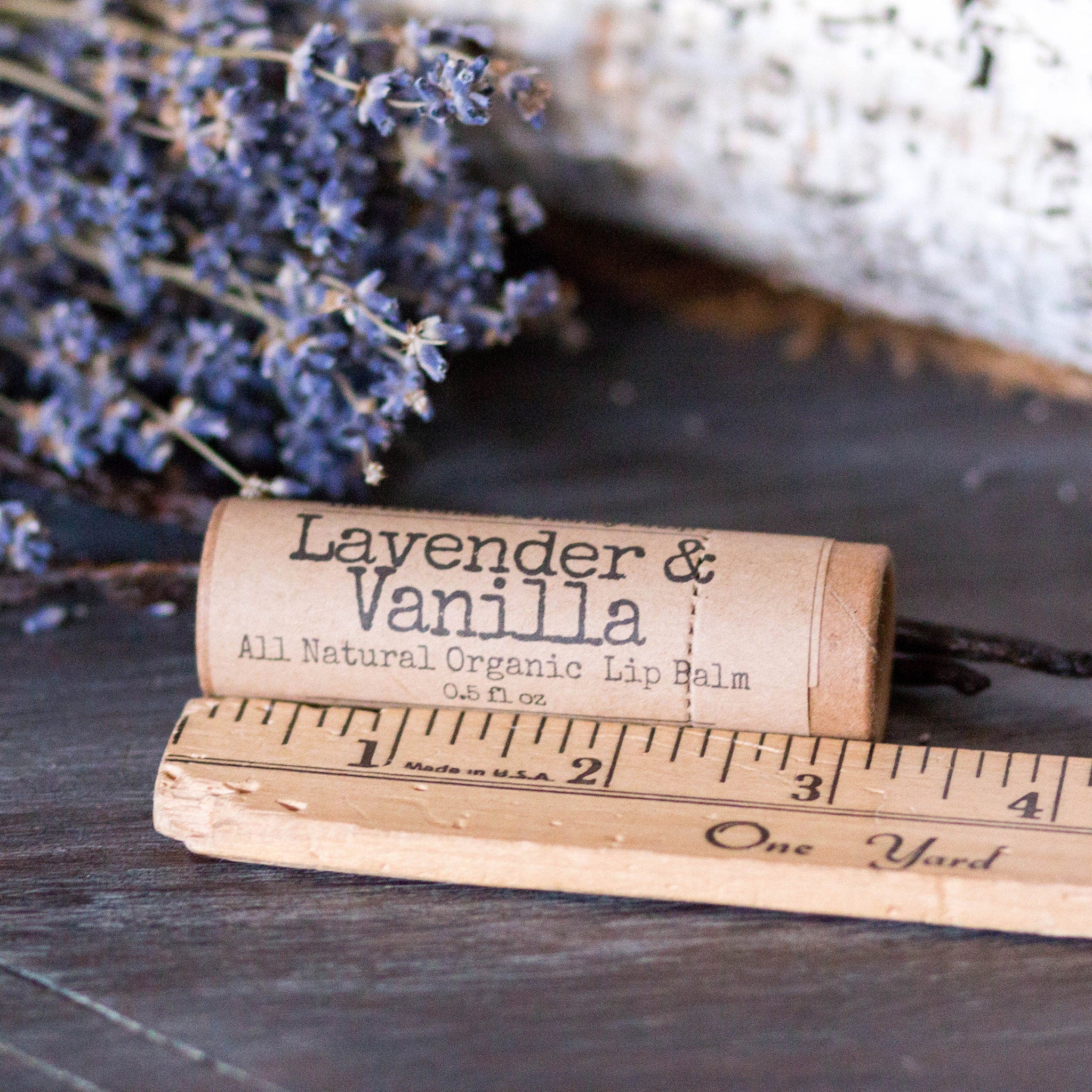 Lip balm tube with "Lavender & Vanilla" text, brown paper, and wooden ruler on dark surface
