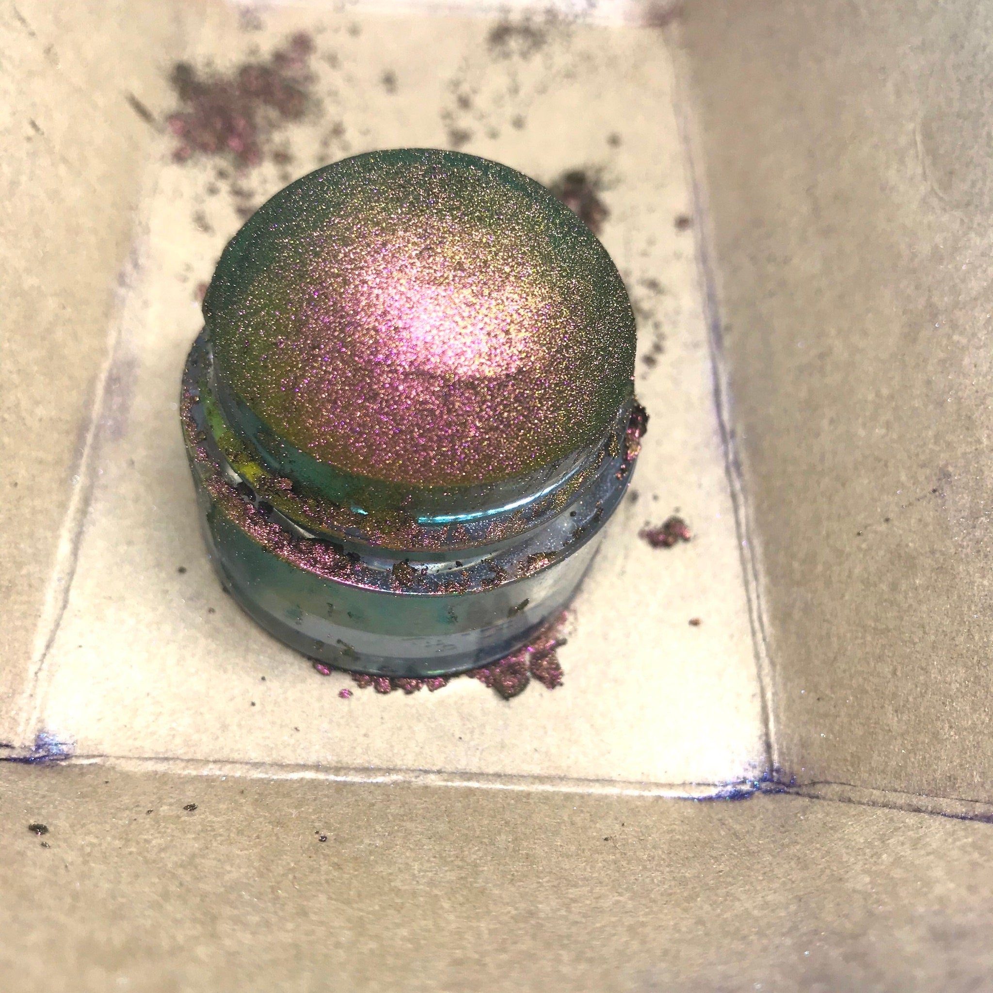 Glitter eyeshadow jar with green, pink, and purple shimmer on a cardboard surface