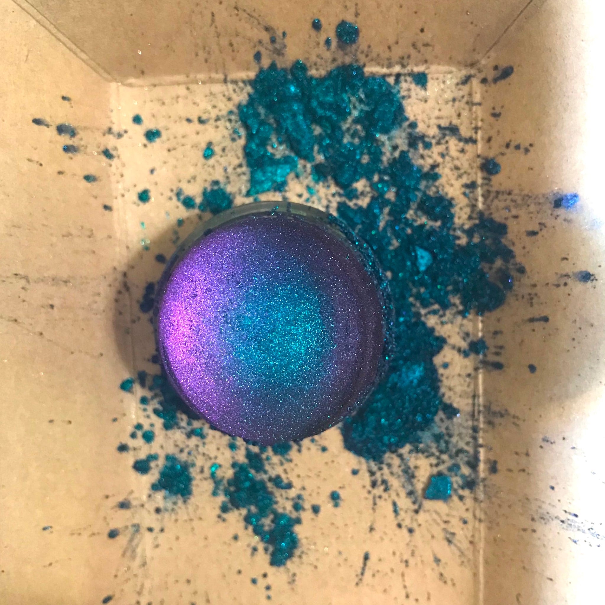 Glitter eyeshadow compact with purple and teal shimmer, spilled glitter on cardboard