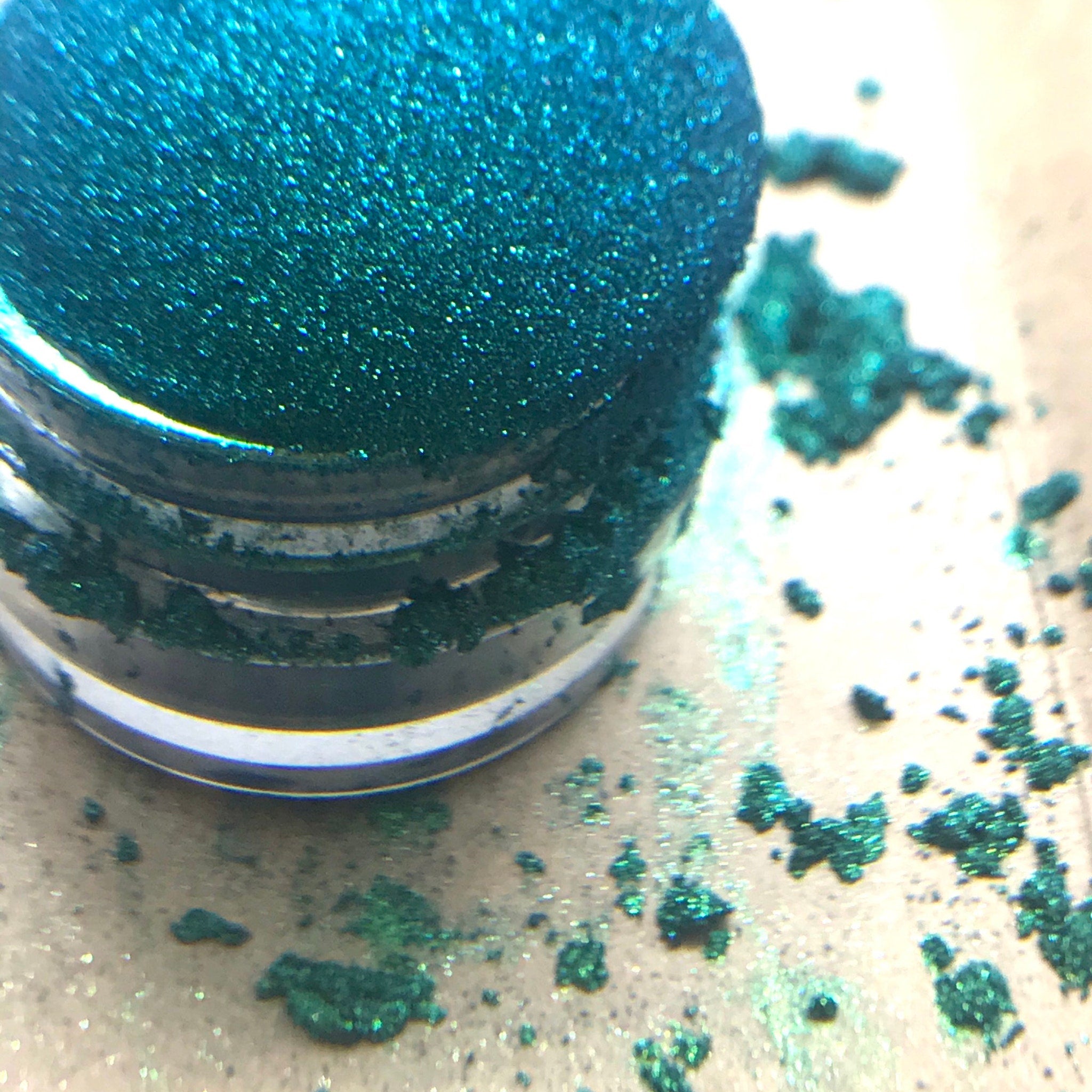 Glitter eyeshadow, teal glitter, small jar, and scattered glitter on a surface