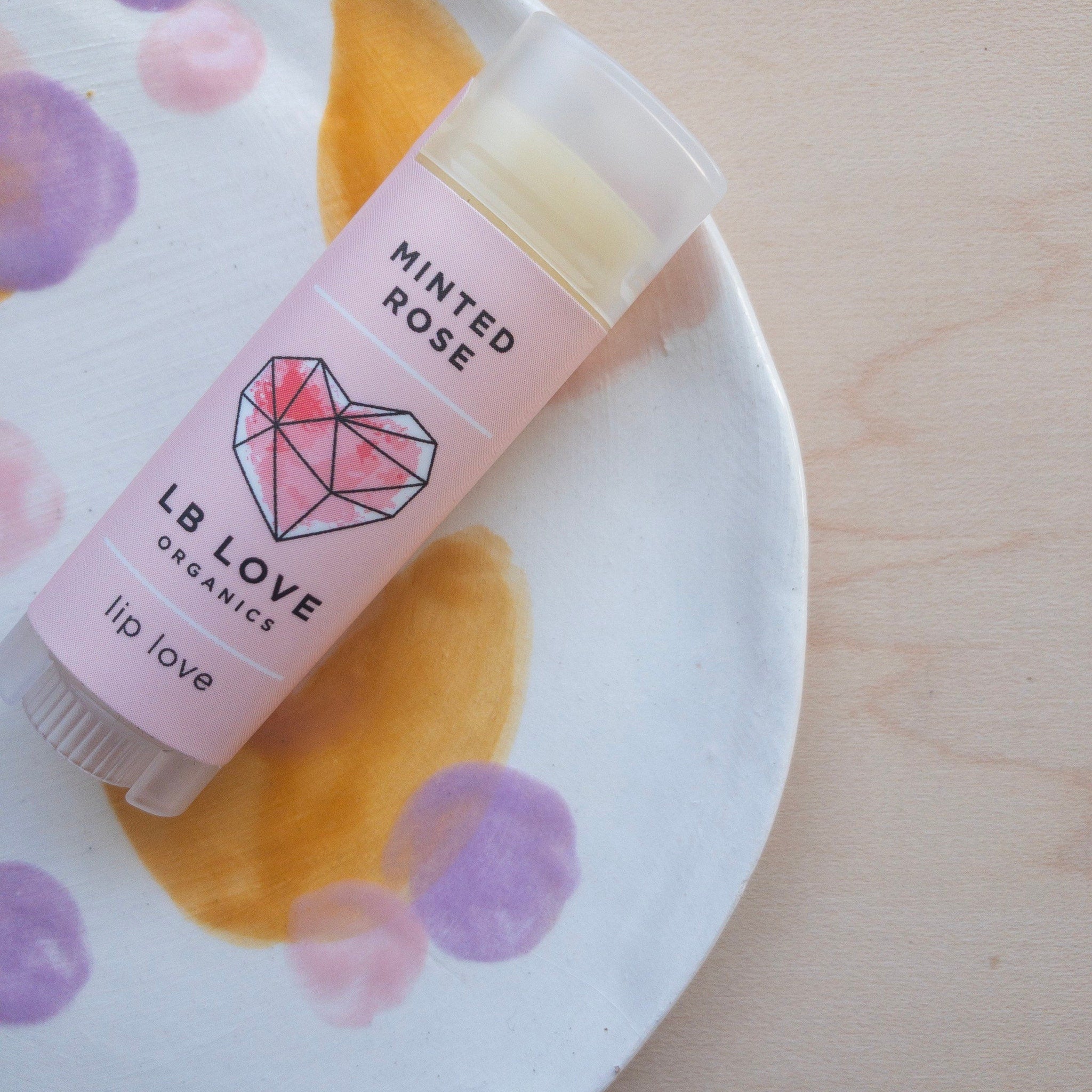 Lip balm tube with pink label, clear tube, and LB LOVE logo on a painted plate