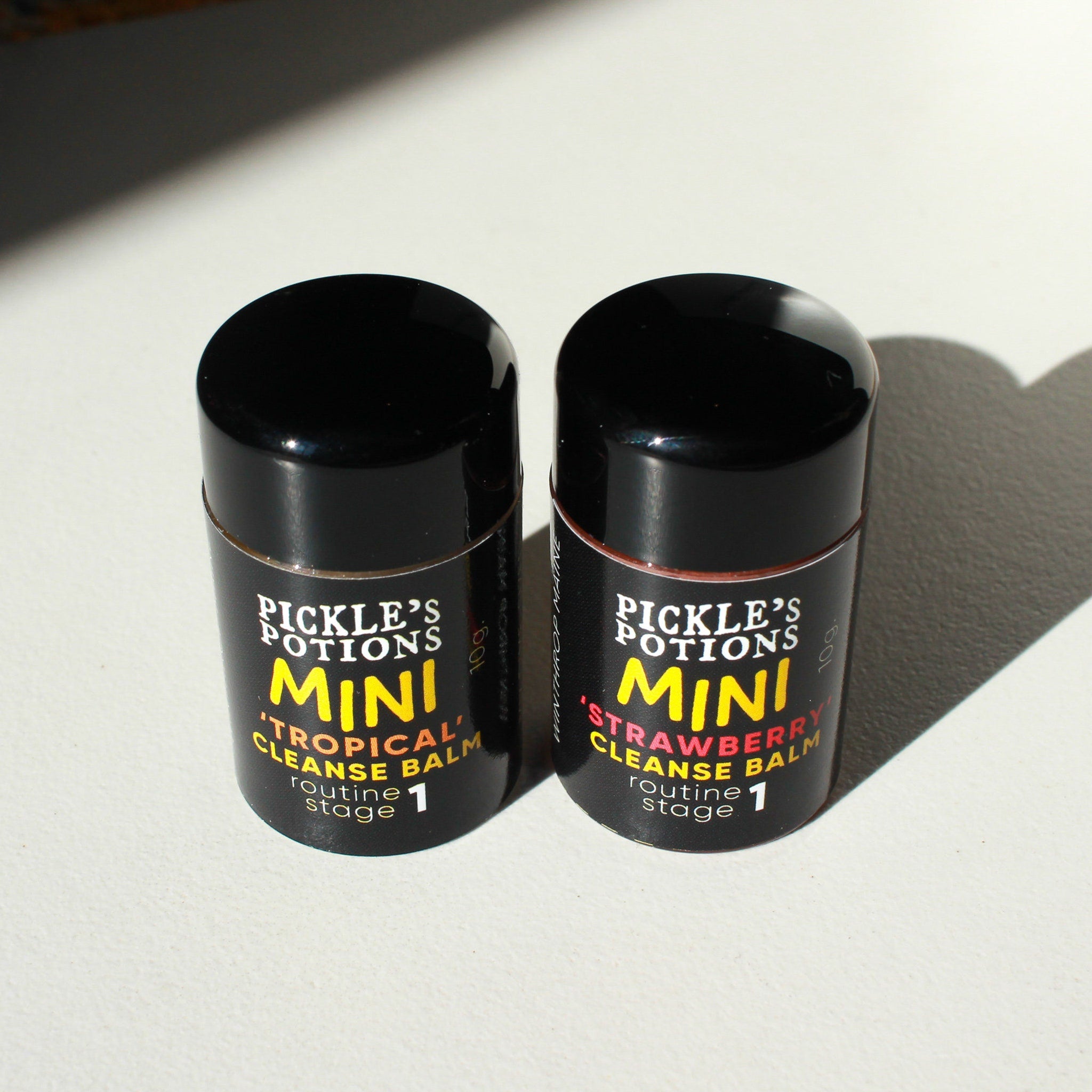 Cleansing balm bottles, black cylindrical containers with yellow and white text, "Pickle's Potions" brand visible, on white surface