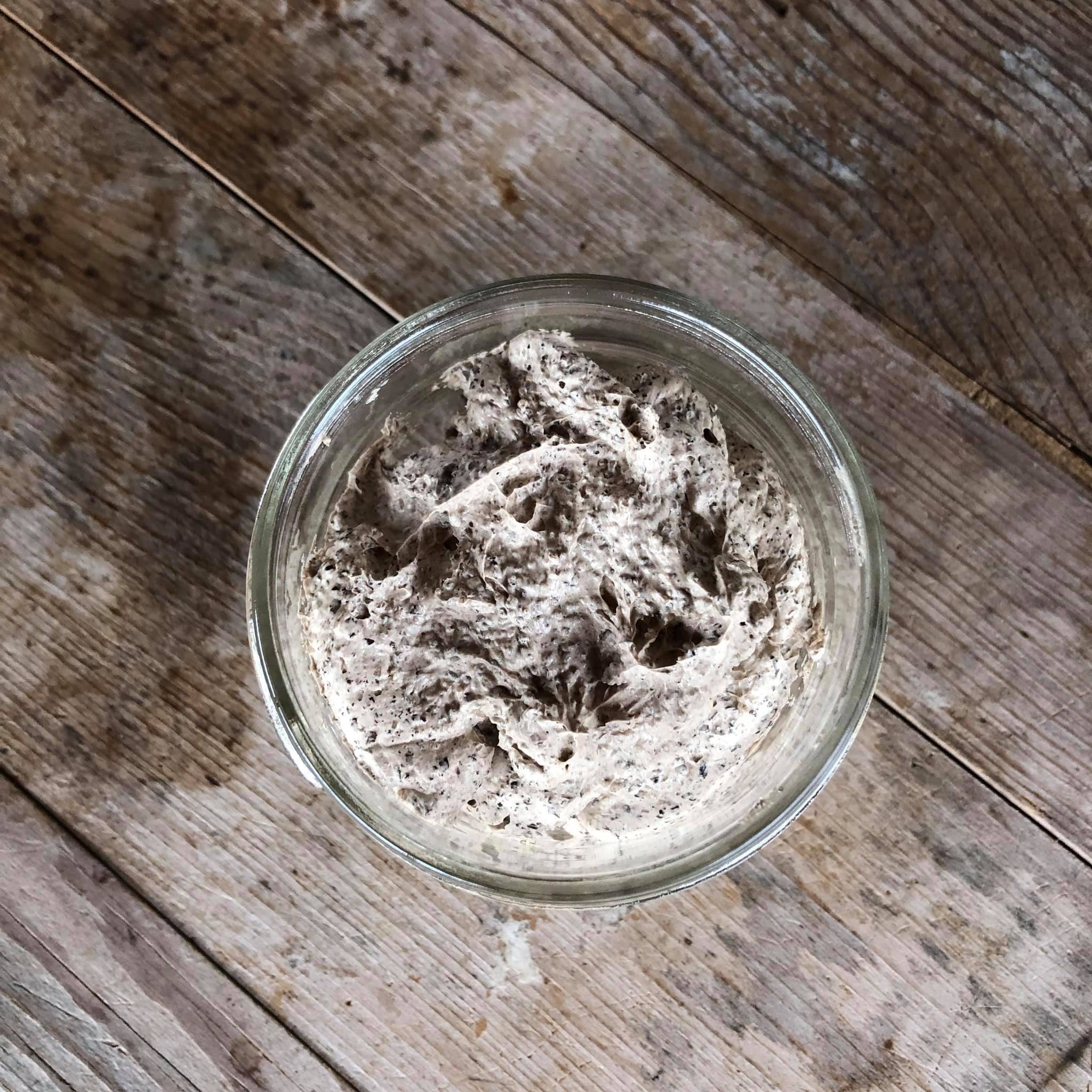 Sourdough starter in glass jar, light brown color with dark specks, on wooden surface