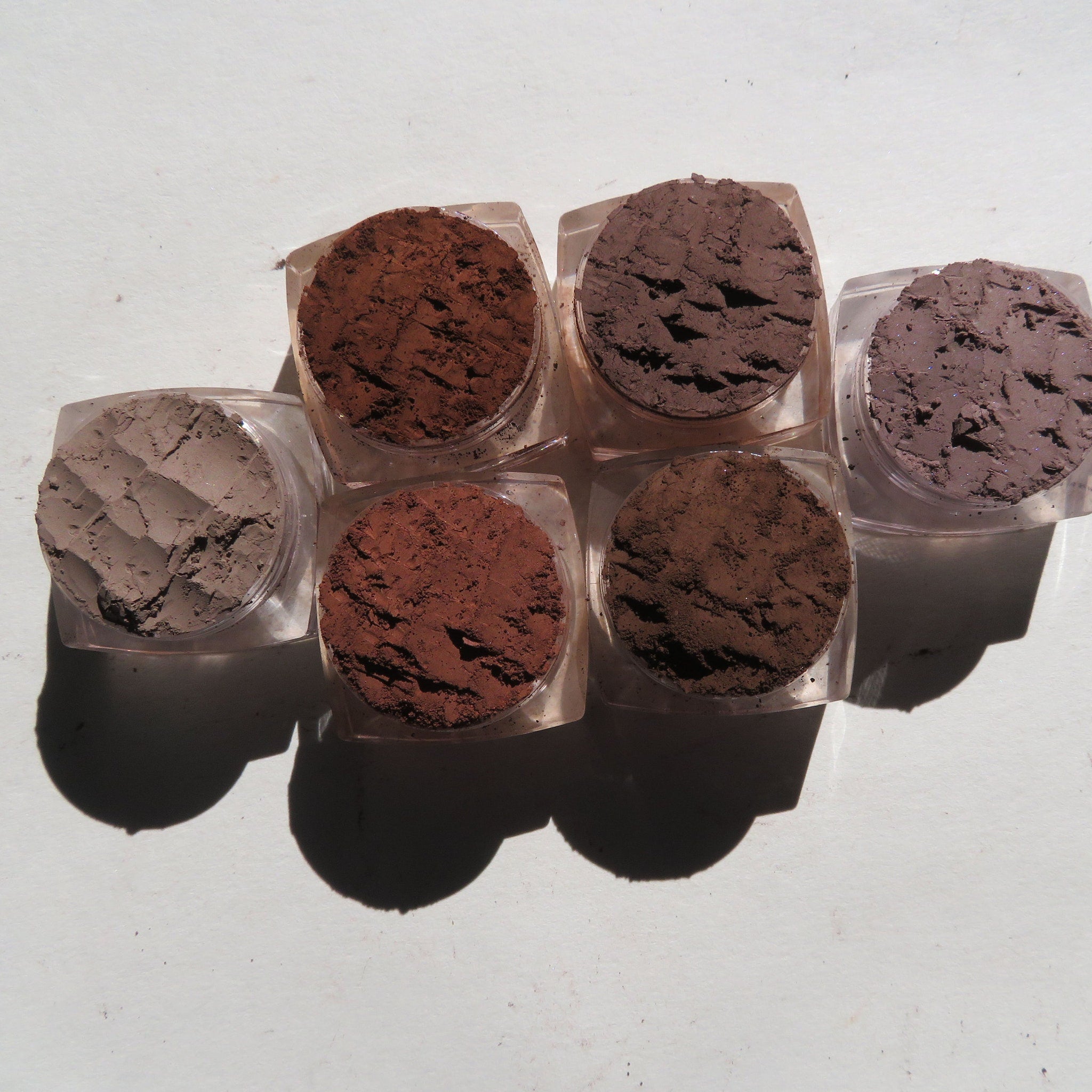 Six matte brown vegan eyeshadow pigments in round jars displayed on a wooden tray with brushes.