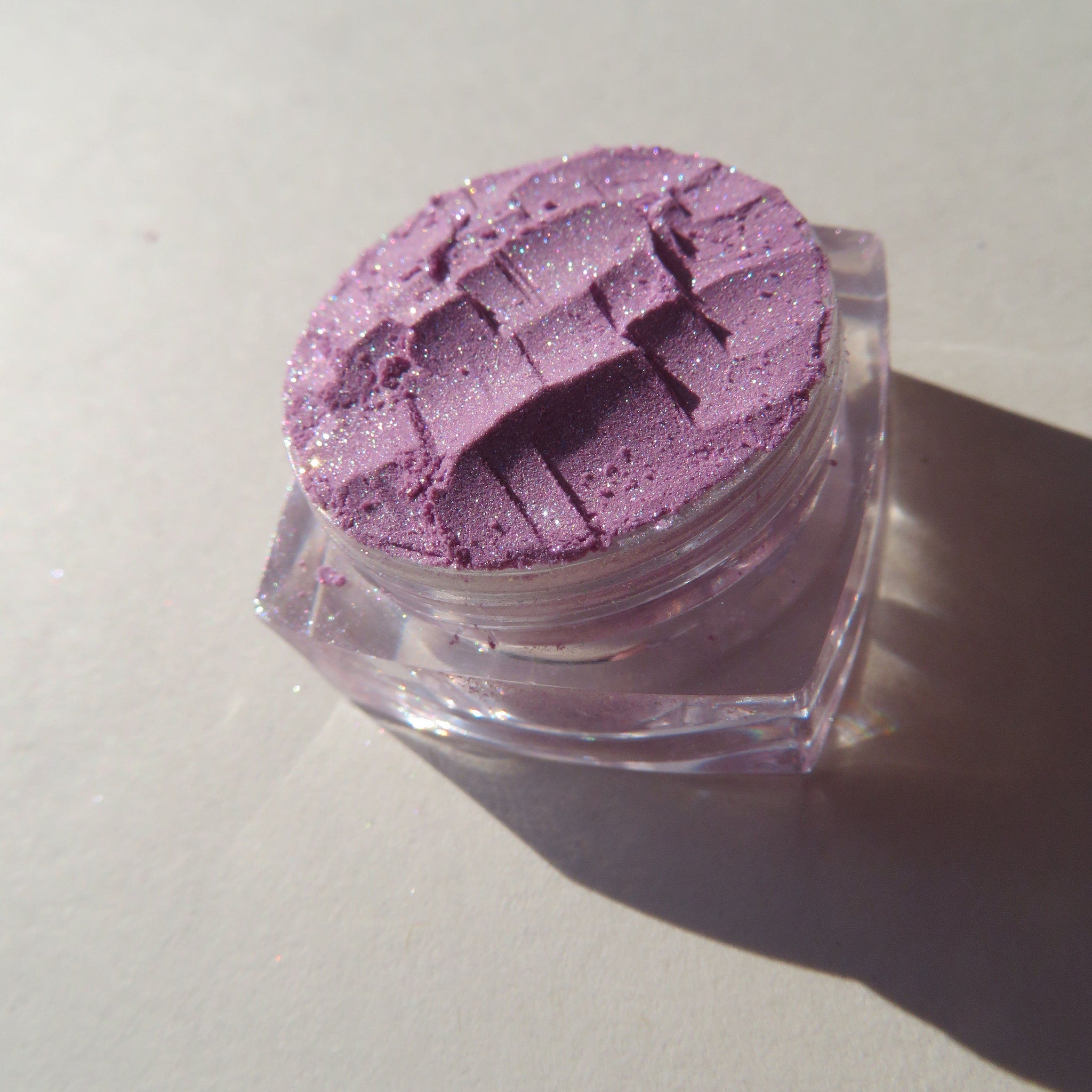Purple glitter eyeshadow with a textured surface in a clear plastic container