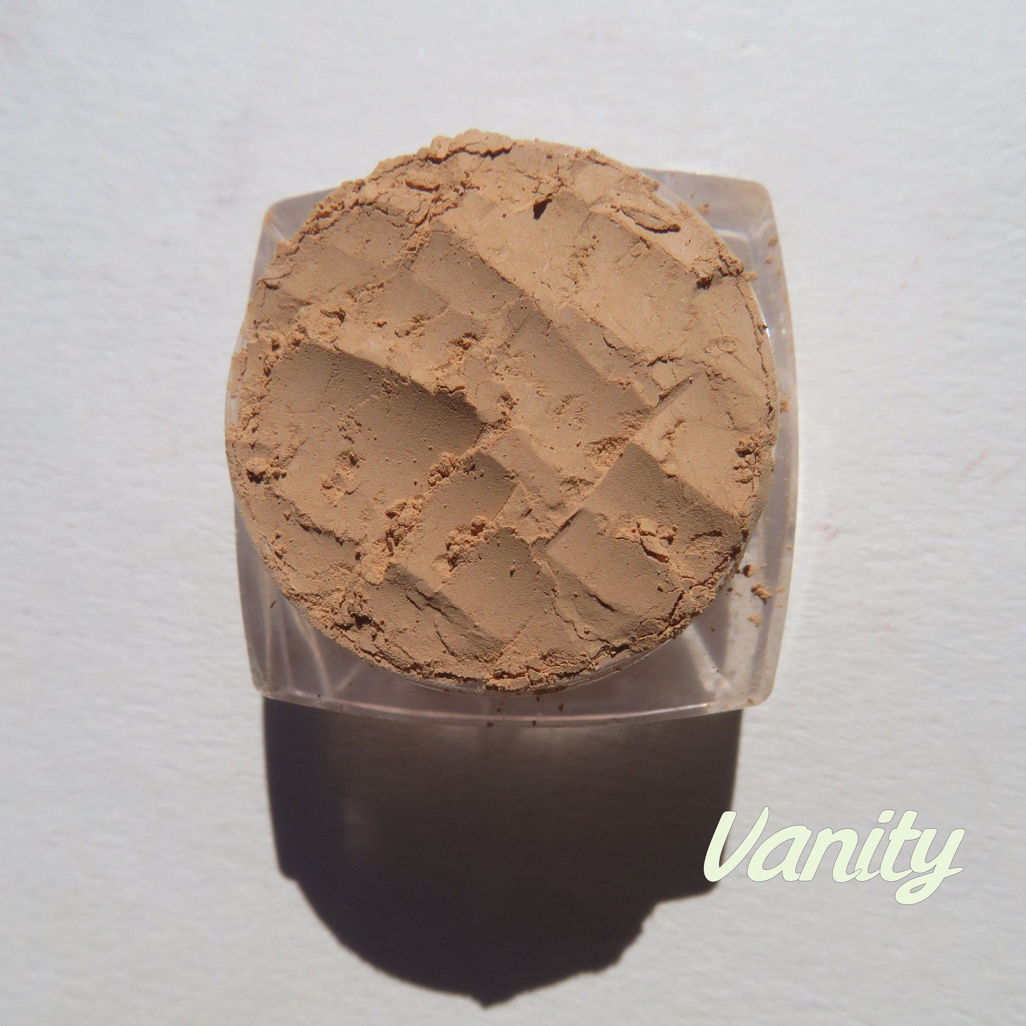 Powder compact with tan powder, clear plastic container, and "Vanity" text on white surface