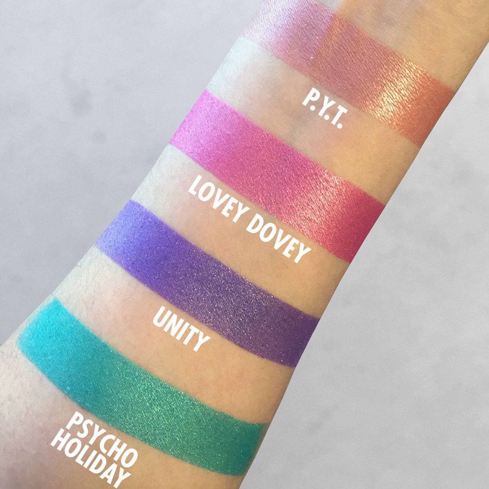 Swatches of eyeshadow in pink, purple, and green with metallic shimmer, names visible on arm