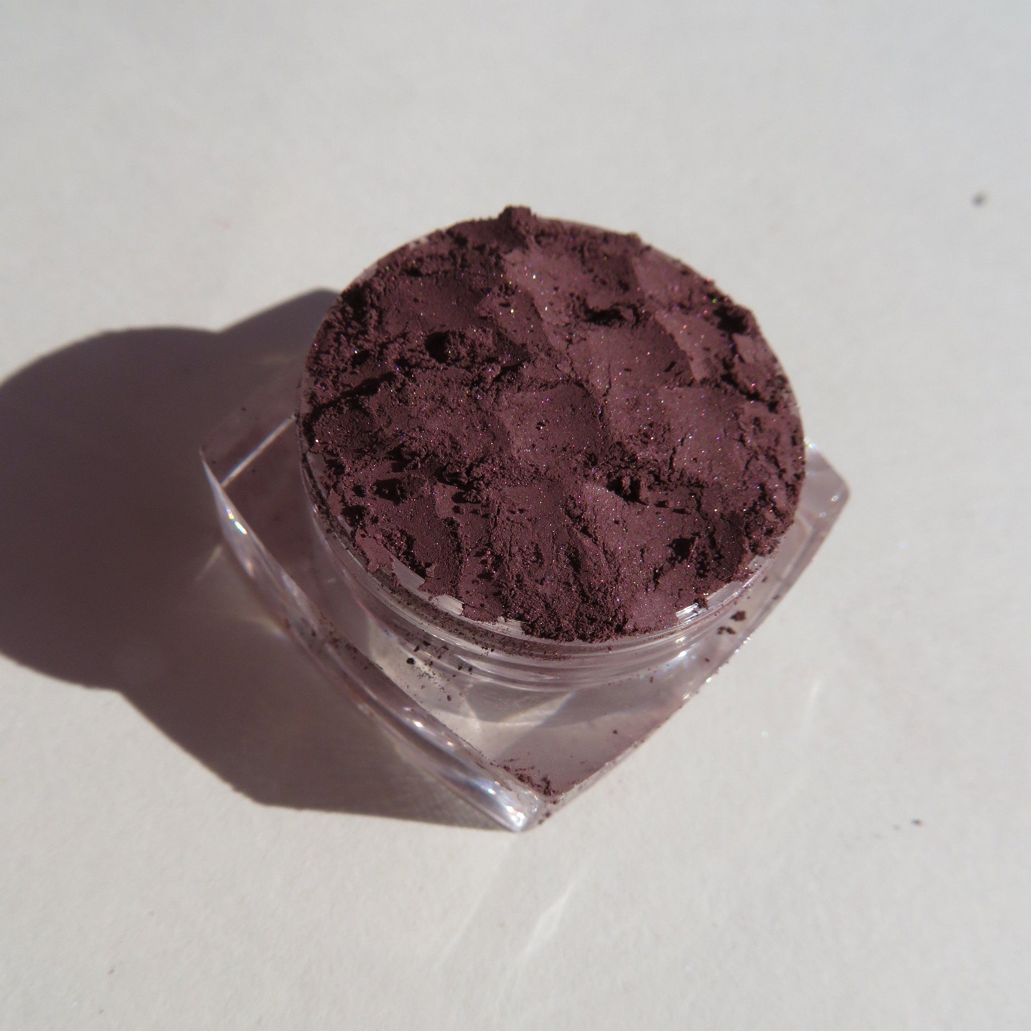 Eyeshadow powder in dark purple with glitter, in clear plastic jar, close-up shot