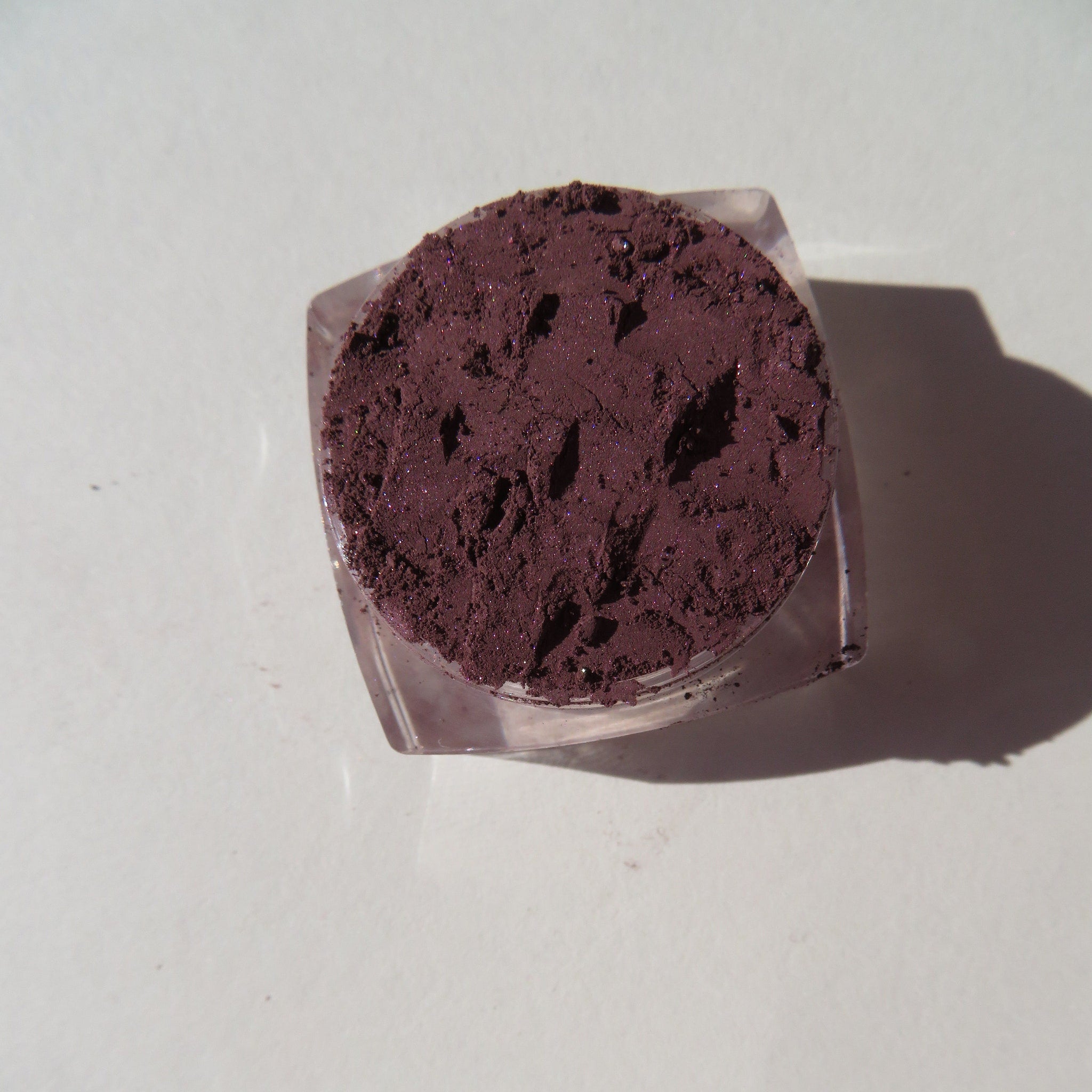 Eyeshadow powder, dark purple with shimmer, in a clear plastic jar, top view