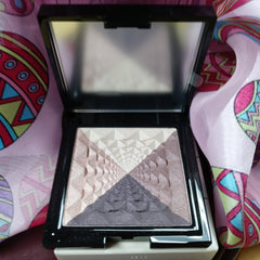 Eye makeup compact with brown and beige eyeshadow colors in a black compact with mirror, resting on patterned fabric
