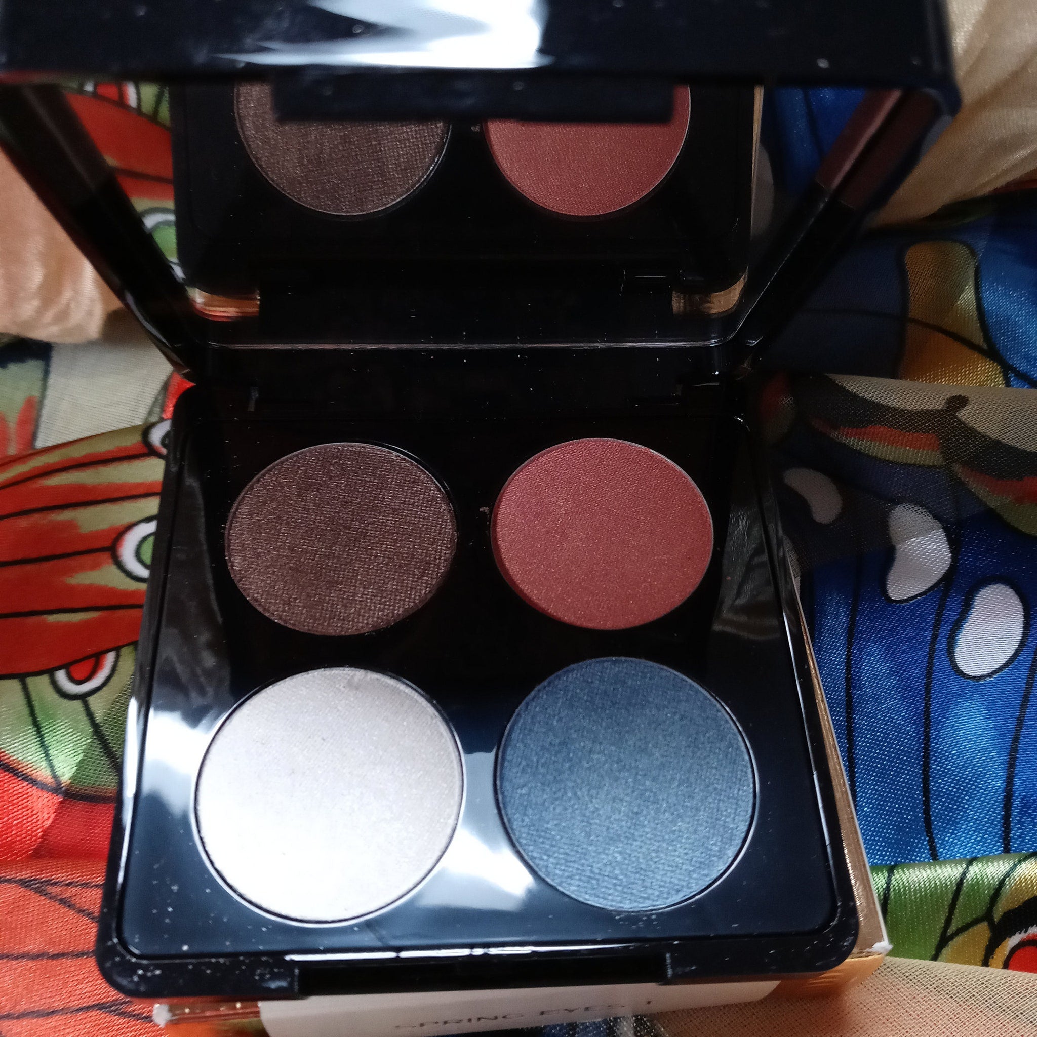 Close-up of four shimmering eyeshadow pans in bronze, plum, taupe, and champagne hues with embossed logo