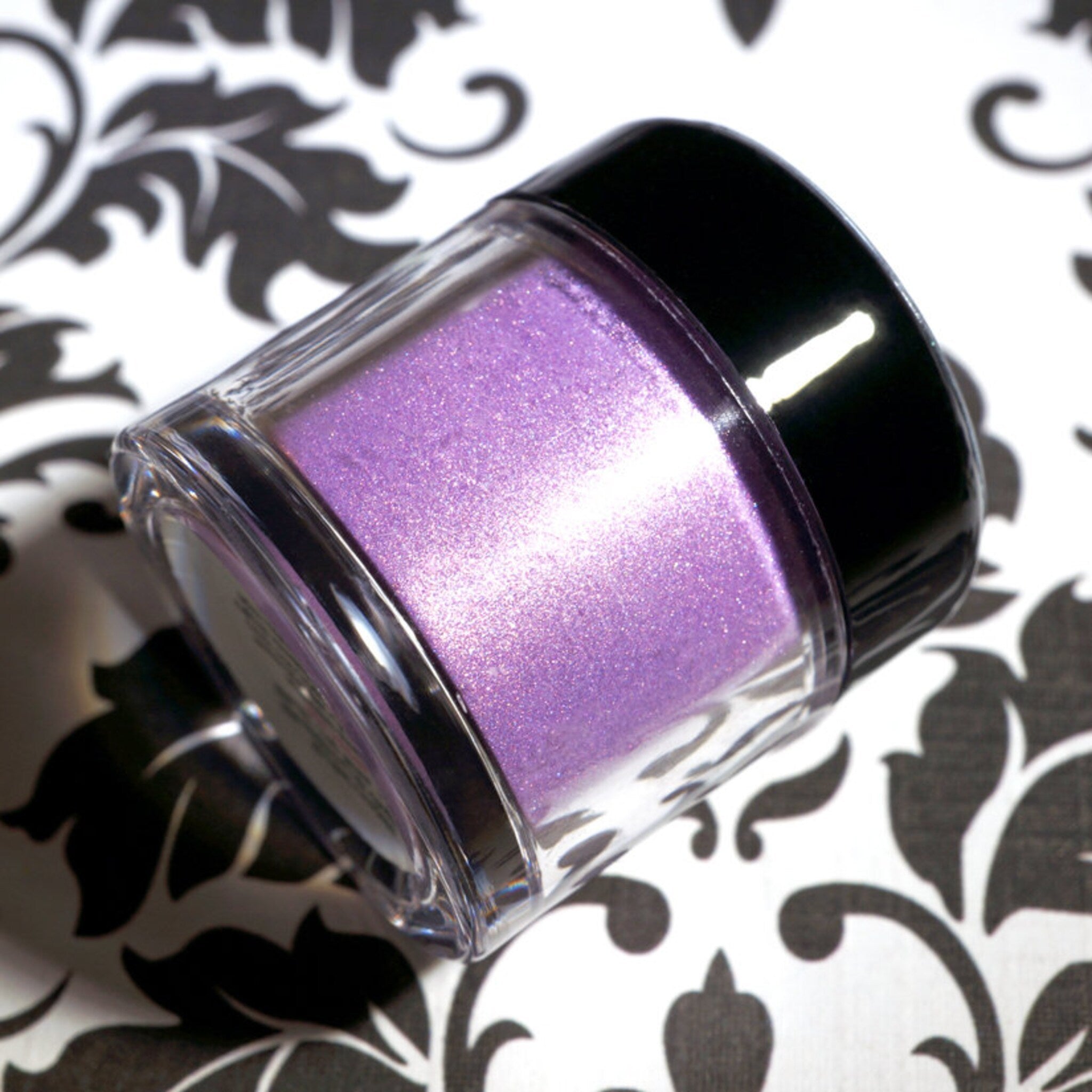 Purple glitter powder in clear glass jar with black cap, on white and black floral pattern