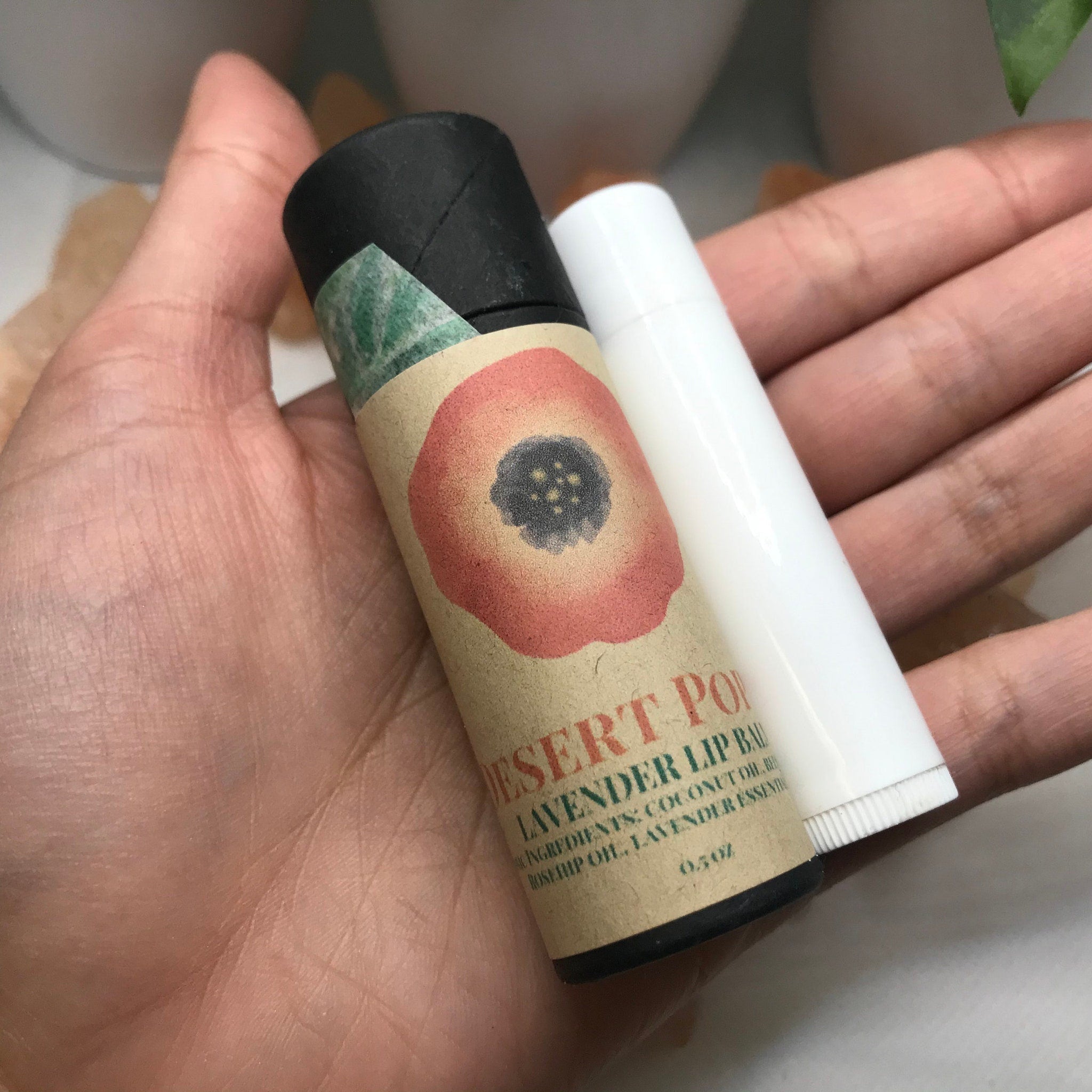Lip balm with black, white, and floral packaging, held in hand, "Desert Po" text visible