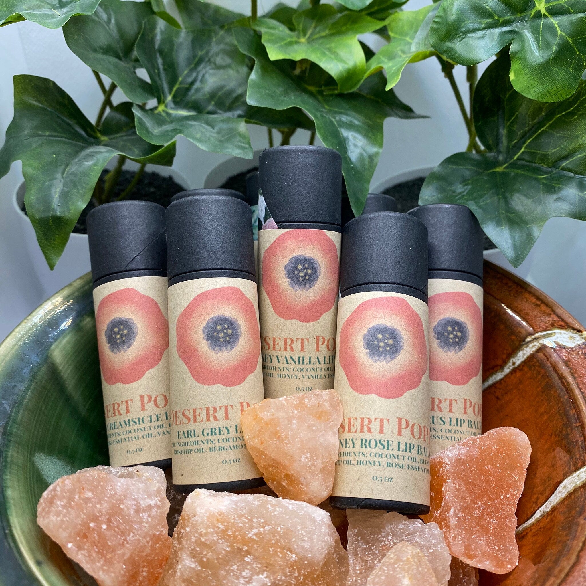 Lip balm tubes with black caps and floral labels, displayed in a bowl with pink salt rocks